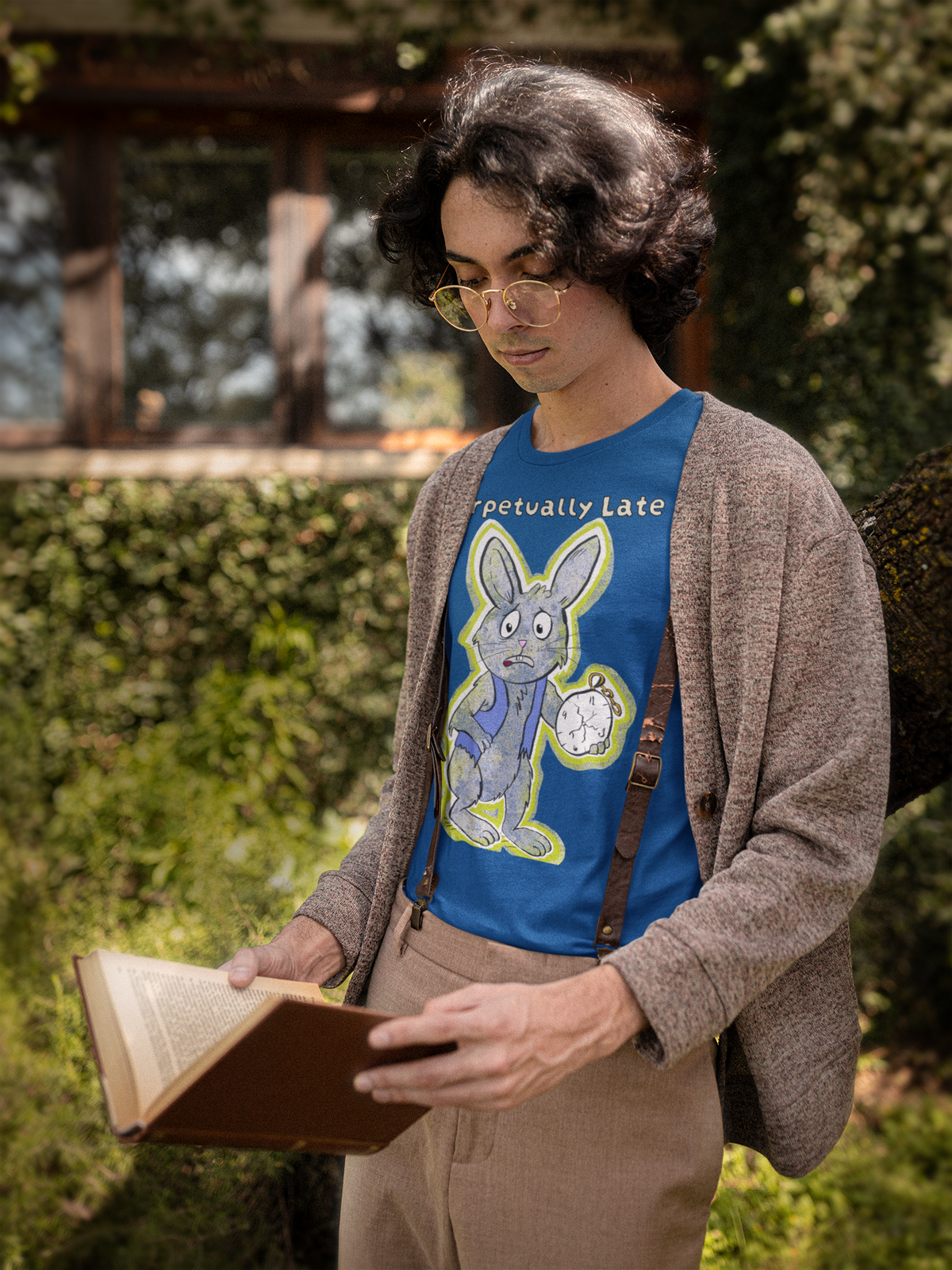 Young man in glasses reading a book, wearing a true royal blue White Rabbit Perpetually Late t-shirt under a brown cardigan. GrungePup That Girl, Alice.