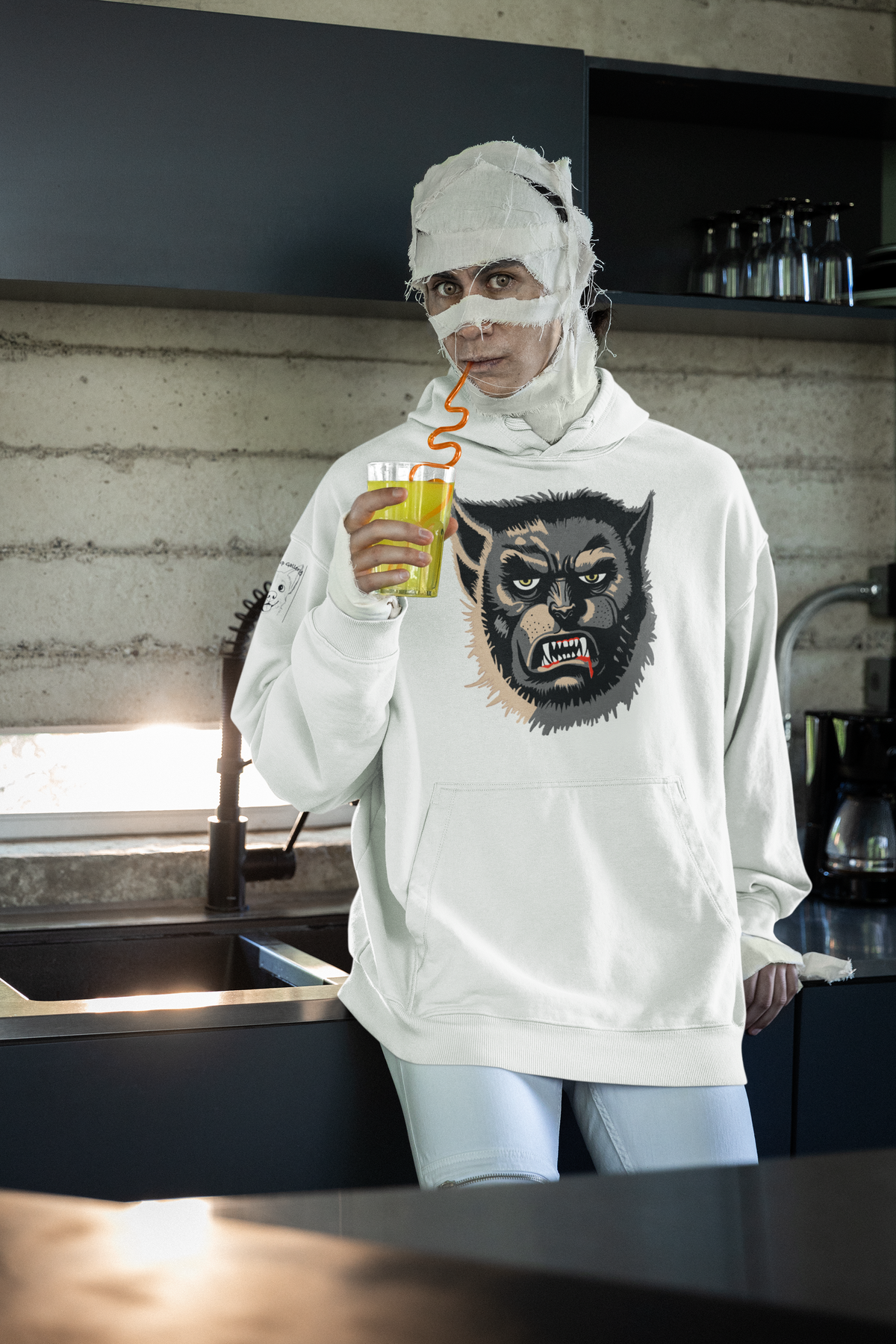 Person wrapped as a mummy drinking juice in a kitchen, wearing a white Wolfman hoodie with the snarling Wolfman face print