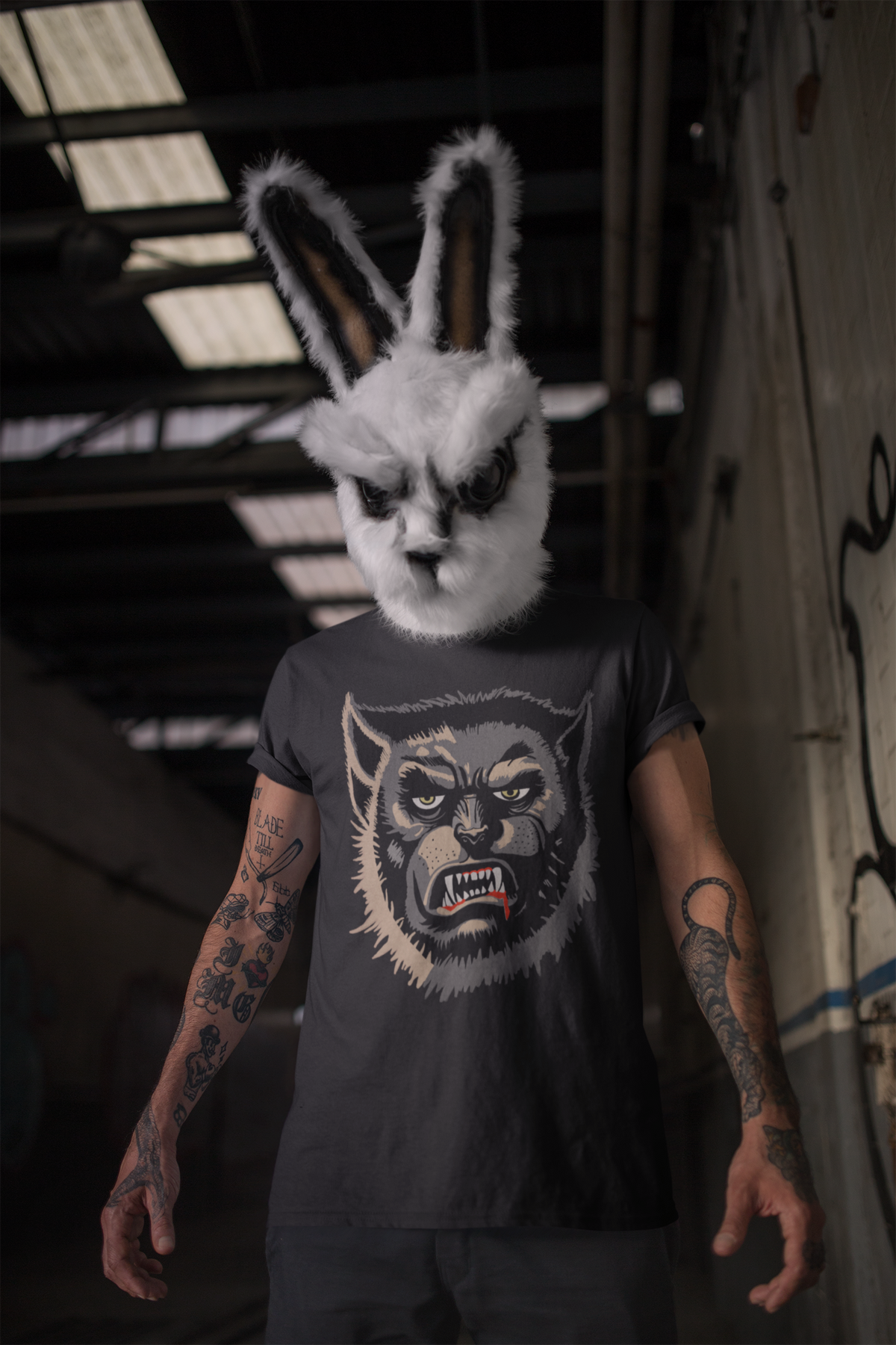 Tattooed man in a white rabbit mask standing in a warehouse, wearing a black Wolfman t-shirt with yellow eyes and bared teeth
