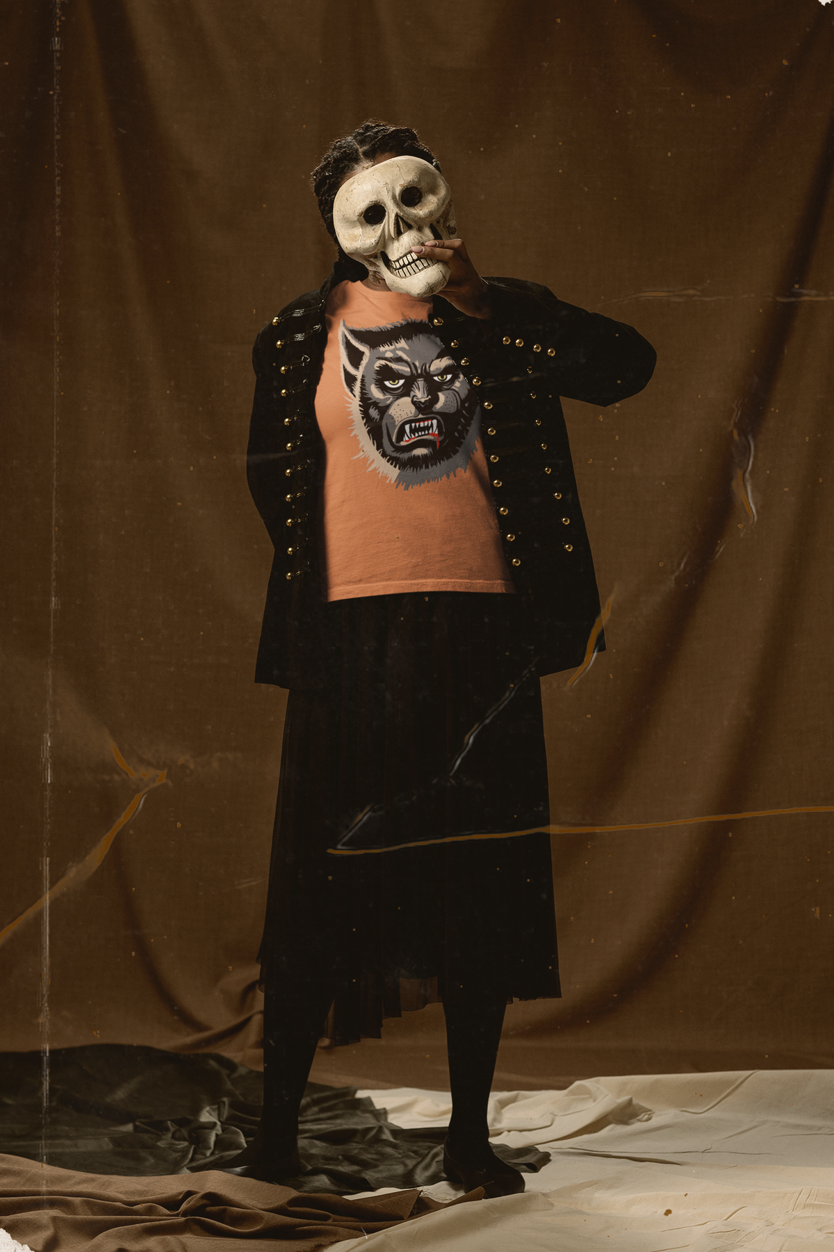 Woman holding a skull mask over her face in a studio, wearing a burnt orange Wolfman t-shirt under a black gold-button jacket