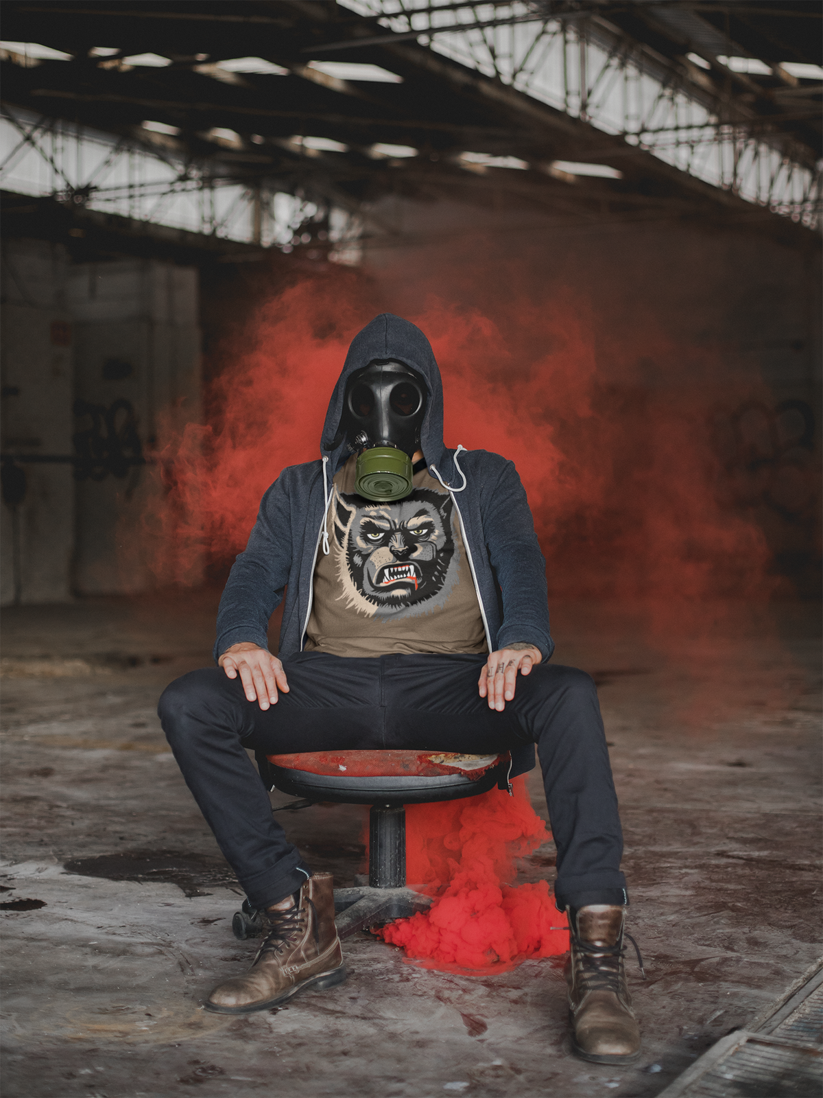 Man in a gas mask and unzipped hoodie sitting in a warehouse with red smoke, wearing a heather brown Wolfman t-shirt