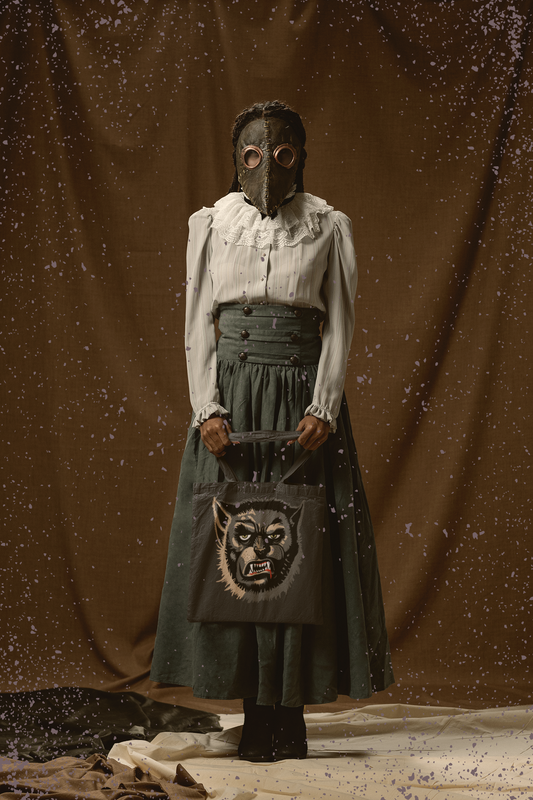 Woman in a vintage dress and plague-doctor mask holding the charcoal Wolfman tote bag, face-only Wolfman print with yellow eyes and bared teeth facing forward