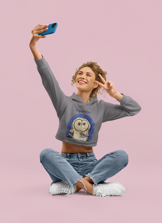 Woman sitting cross legged taking a selfie while wearing a storm blue Bella + Canvas cropped hoodie featuring the Aquarius owl design
