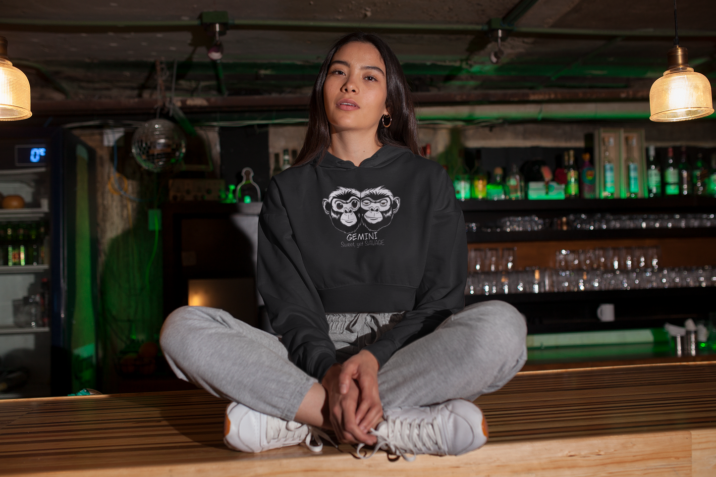 Woman sitting cross legged in Indian style wearing a black Bella + Canvas cropped hoodie featuring the black and white Gemini monkey design