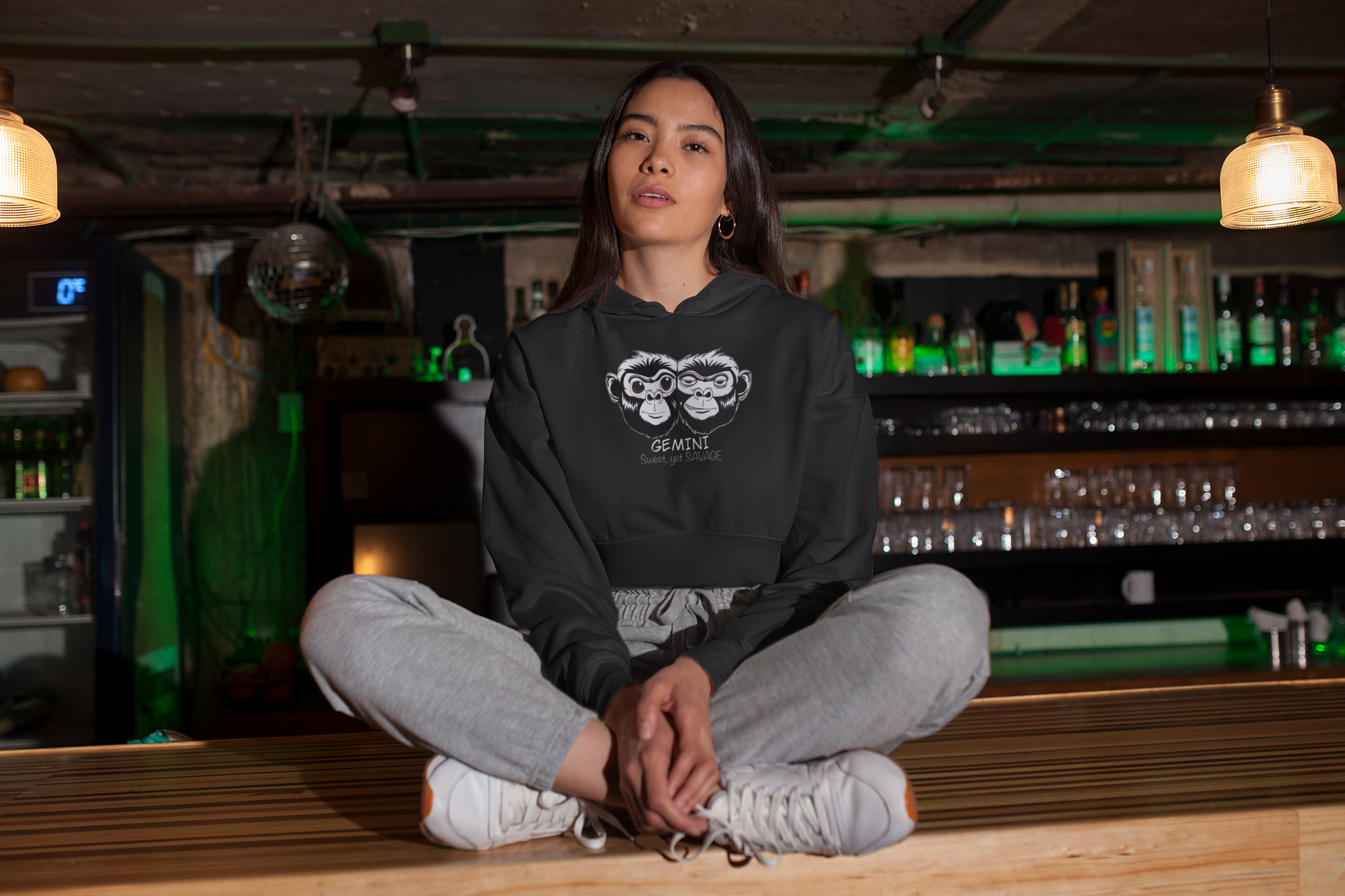 Woman sitting cross legged in Indian style wearing a black Bella + Canvas cropped hoodie featuring the black and white Gemini monkey design