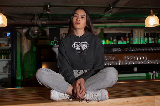Woman sitting cross legged in Indian style wearing a black Bella + Canvas cropped hoodie featuring the black and white Gemini monkey design