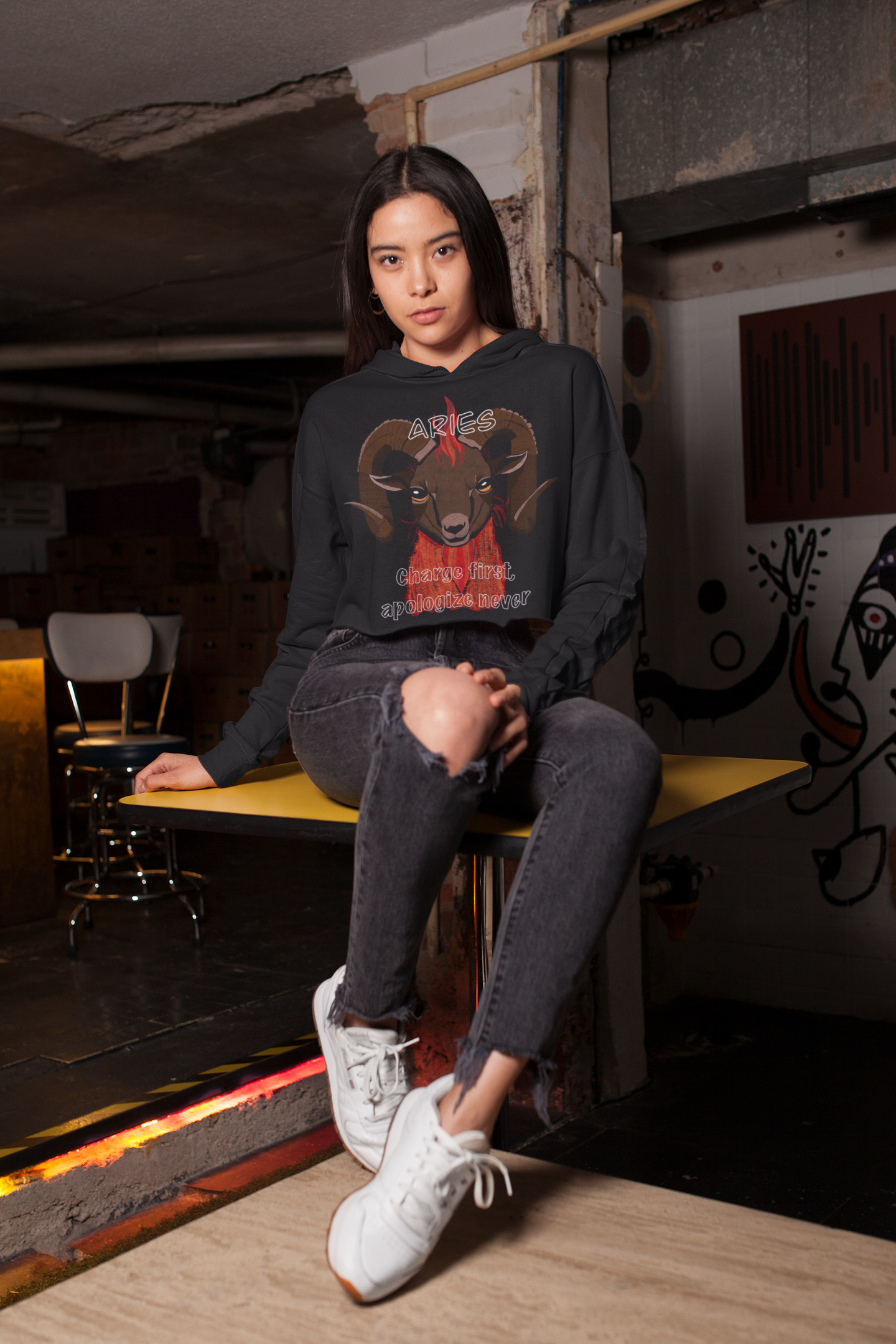 Woman sitting indoors wearing a black cropped hoodie with Aries ram zodiac artwork by GrungePup