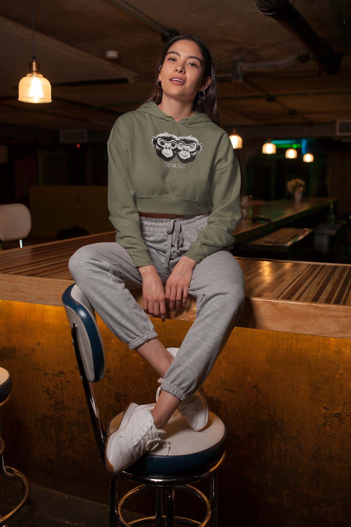 Woman sitting on a bar with legs hanging down wearing a military green cropped hoodie with the Gemini Sweet Yet Savage black and white monkey design