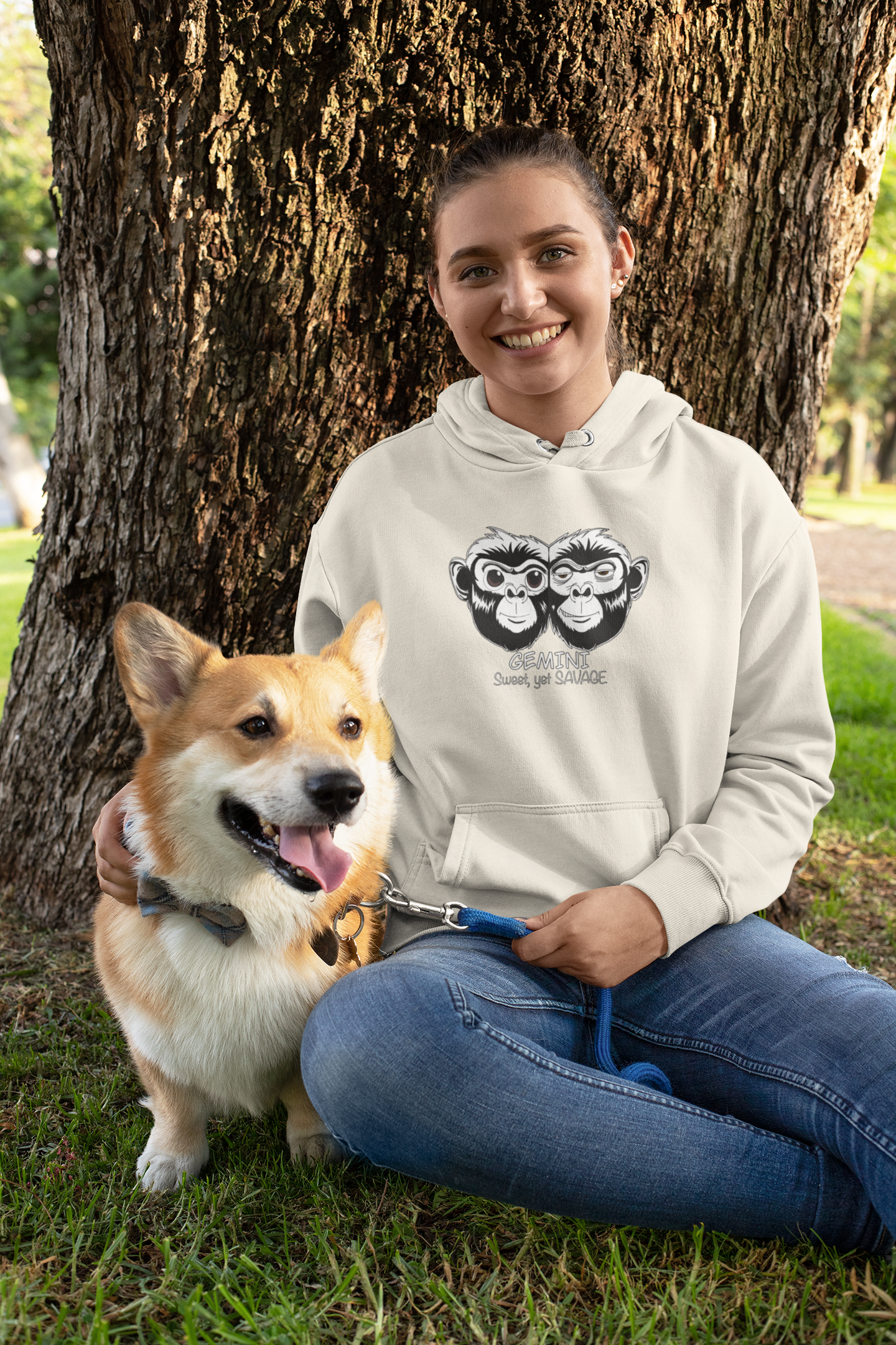 Woman sitting outdoors under a tree with her corgi wearing an oatmeal heather Gemini Sweet Yet Savage hoodie