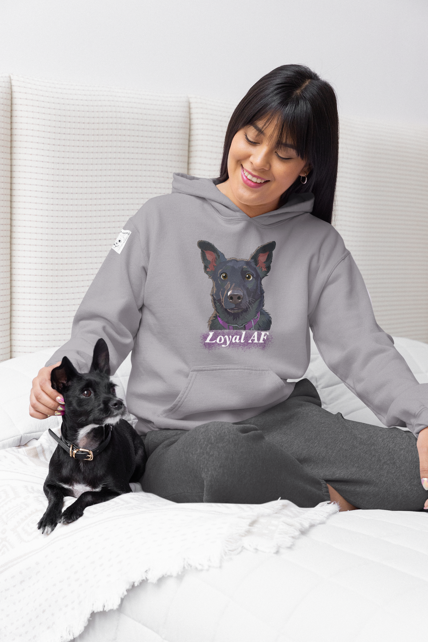 Woman sitting on bed smiling with her small black dog while wearing a sport grey Mathis Loyal AF hoodie