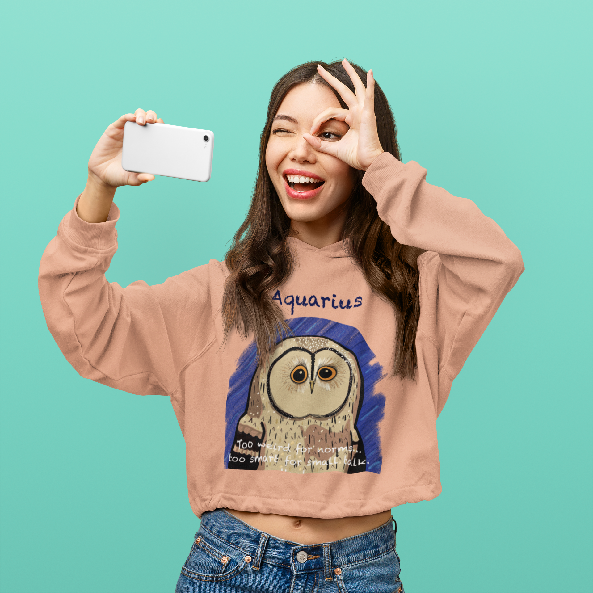 Woman standing and taking a selfie while wearing a peach colored cropped hoodie with the Aquarius owl zodiac illustration