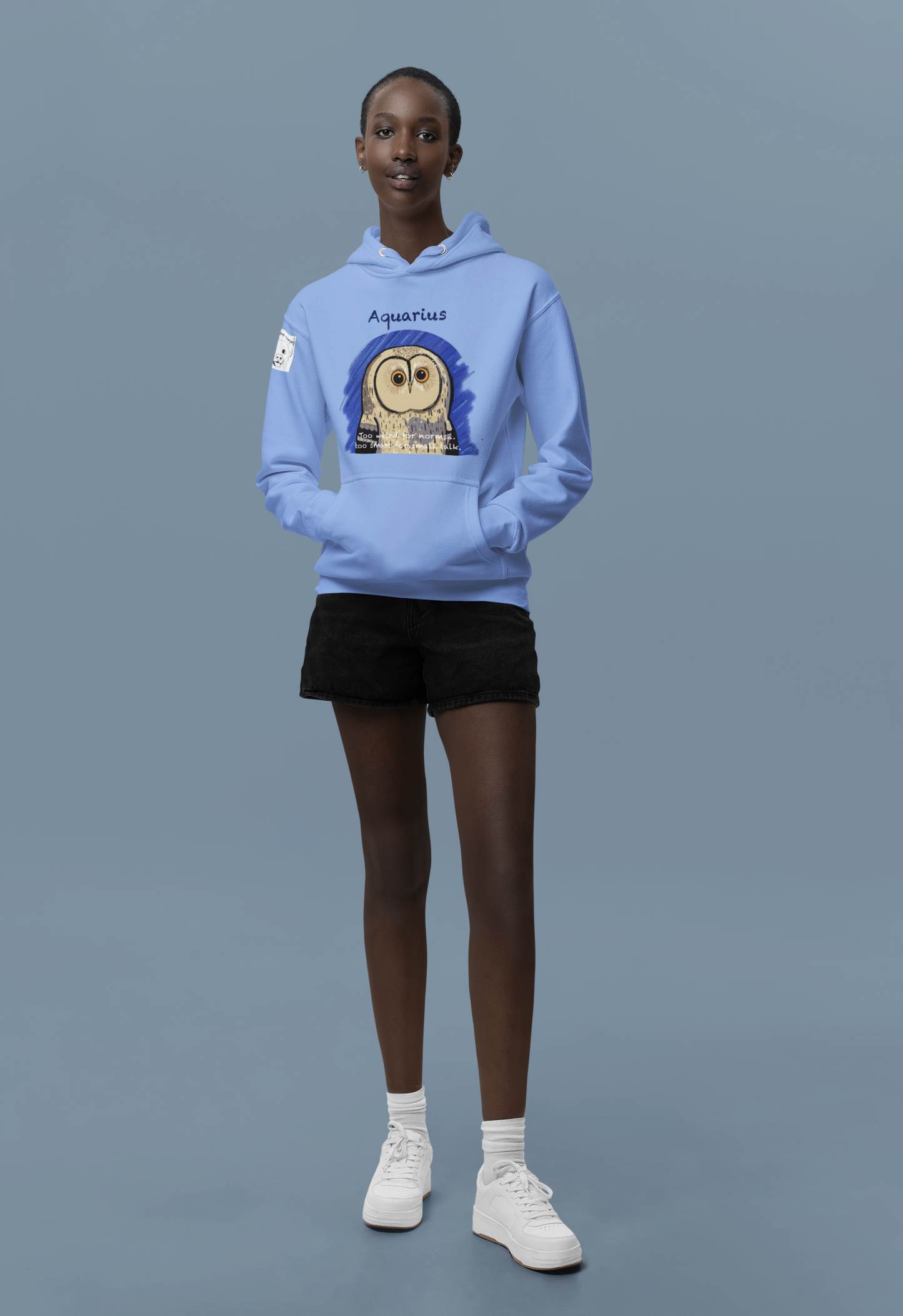 Woman standing in a studio wearing a carolina blue Cotton Heritage hoodie featuring the Aquarius Owl design from GrungePup Sarcastic Zodiac