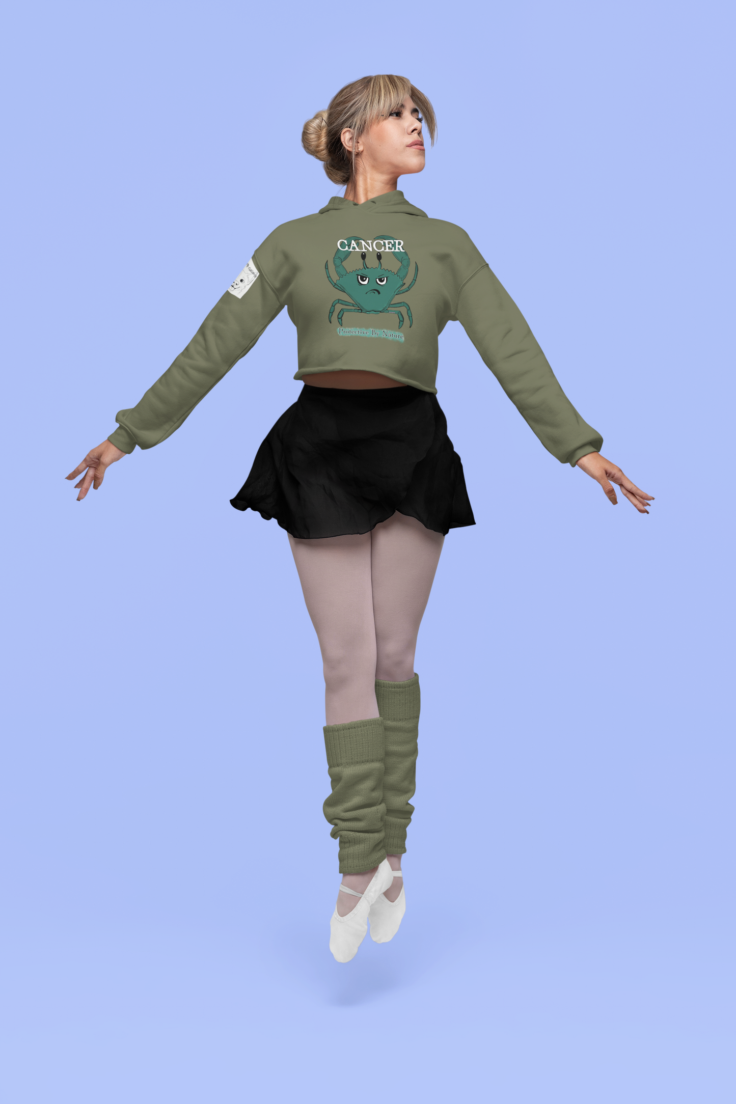 Woman performing a dance pose in a military green Cancer "Protective By Nature" cropped hoodie against a blue studio background