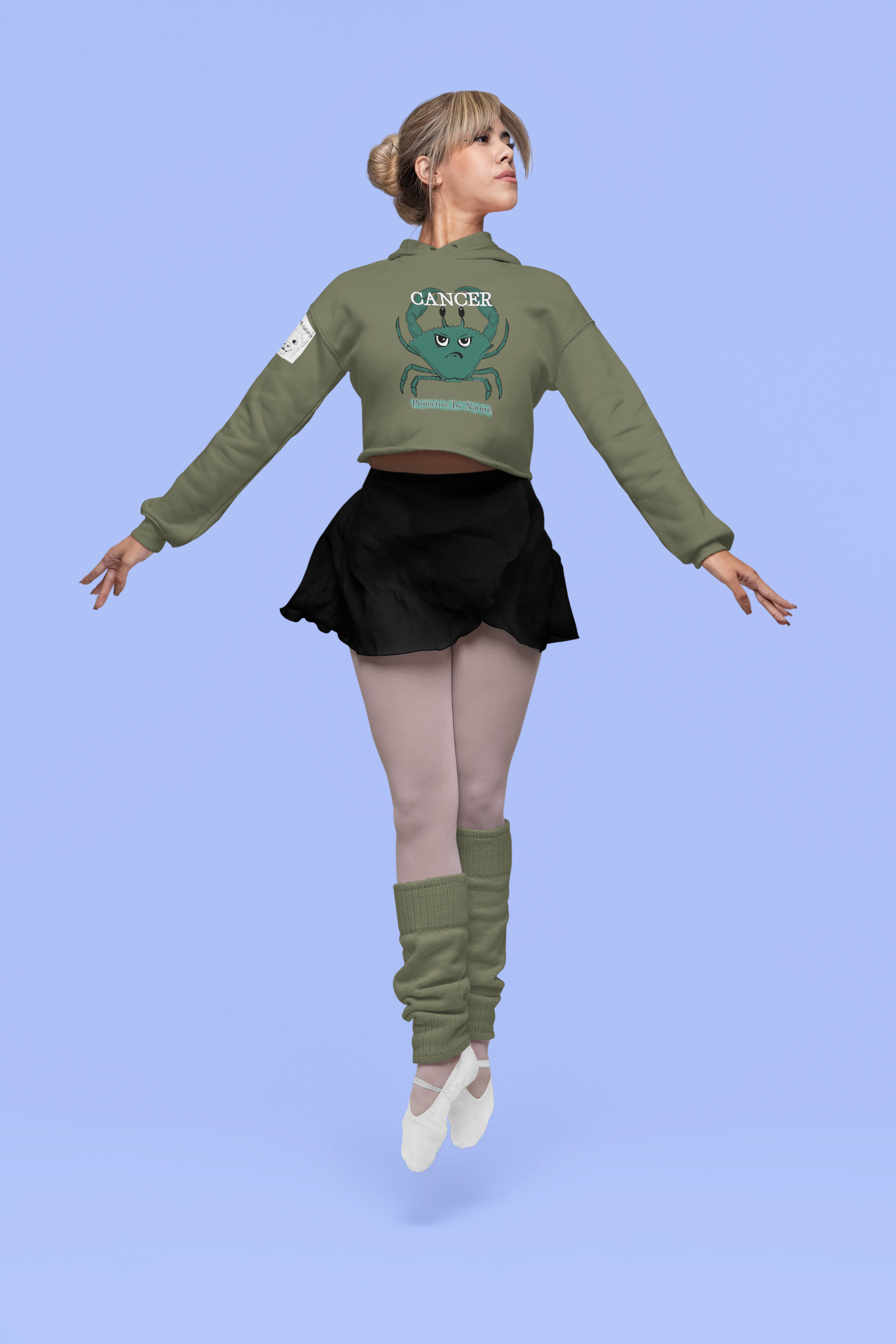 Woman performing a dance pose in a military green Cancer "Protective By Nature" cropped hoodie against a blue studio background