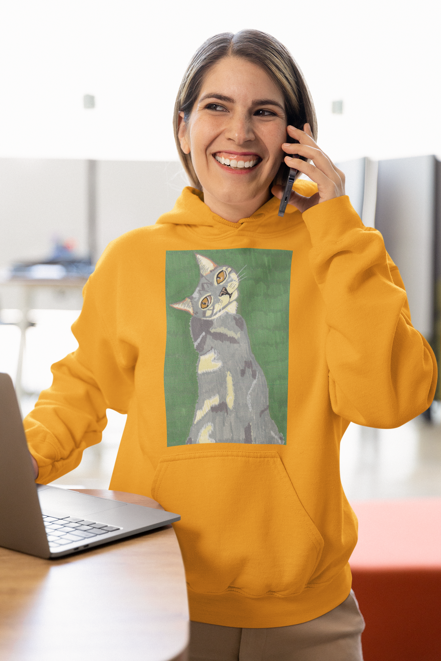 Smiling woman wearing a gold yellow hoodie with Mr. Grey cat portrait while talking on the phone in an office setting