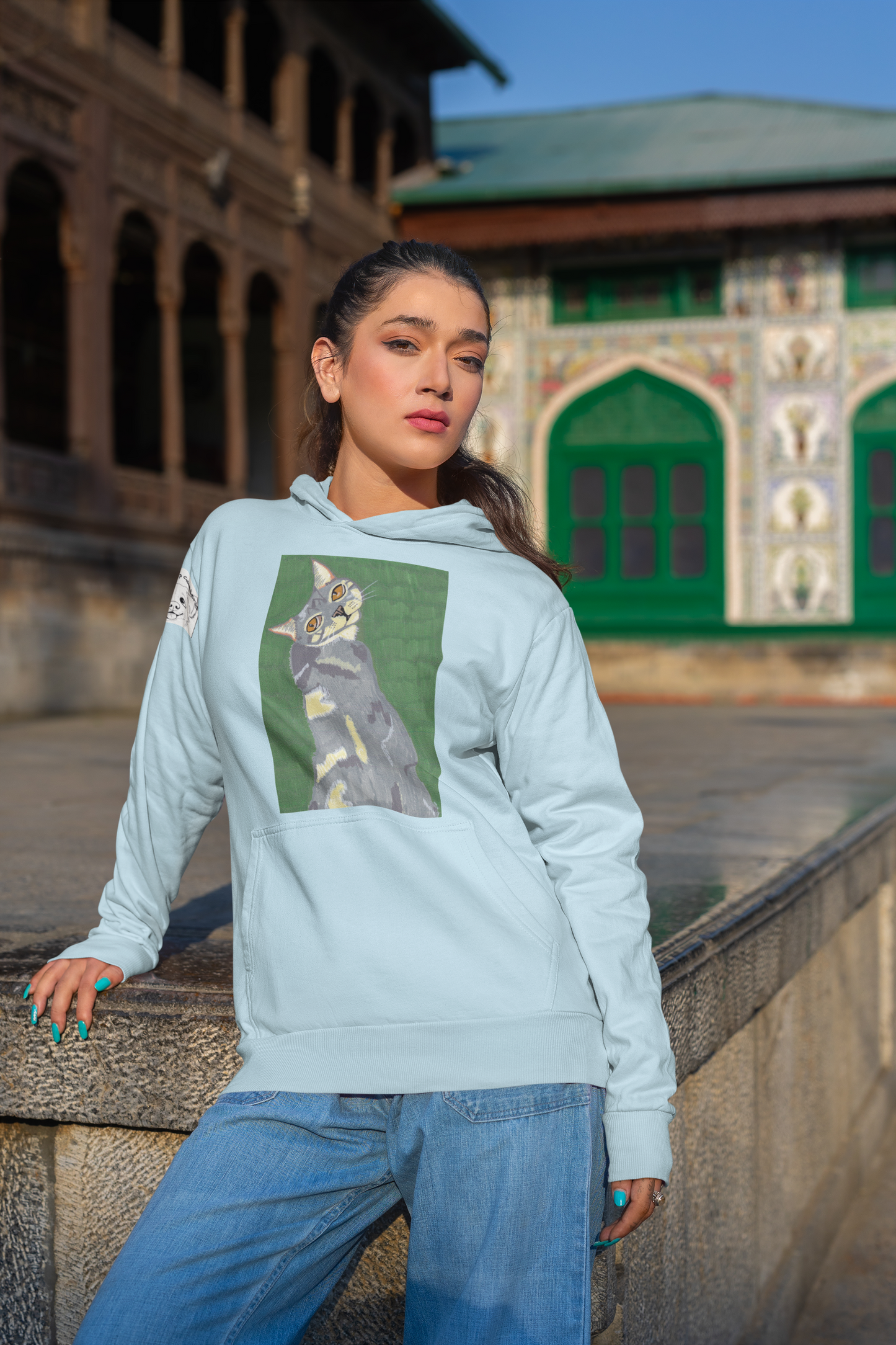 Woman leaning against a stone wall wearing a light blue hoodie featuring the Mr. Grey cat portrait from GrungePup