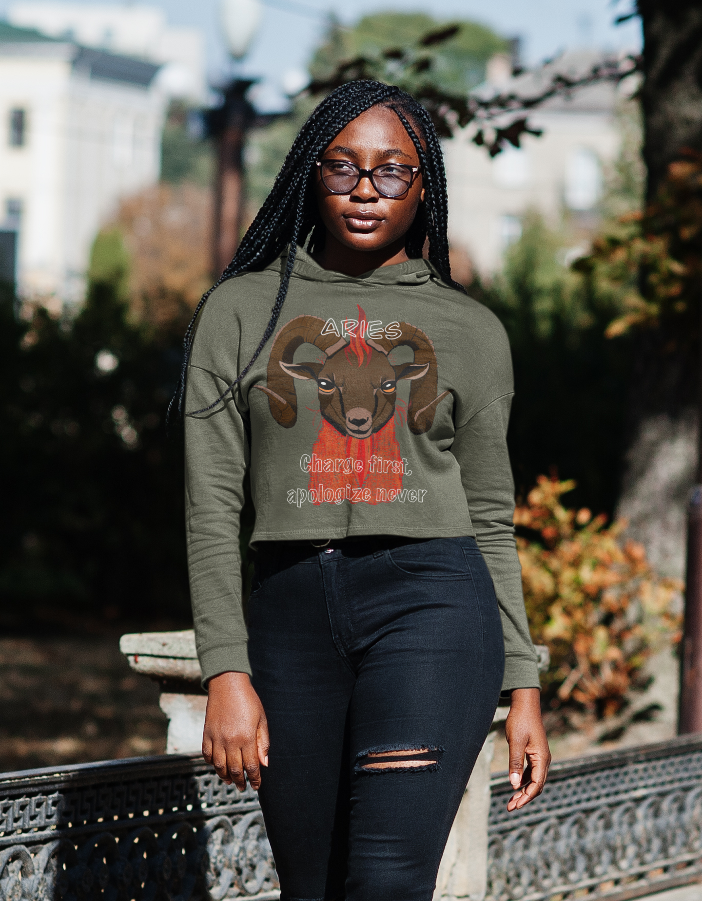 Woman wearing sunglasses walking outdoors in a military green Bella + Canvas cropped hoodie featuring the Aries ram design