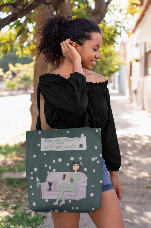 Woman with curly hair wearing black off-shoulder top holding a dark green Feed Me Chocolate and Call Me Pretty tote bag on a sunny street
