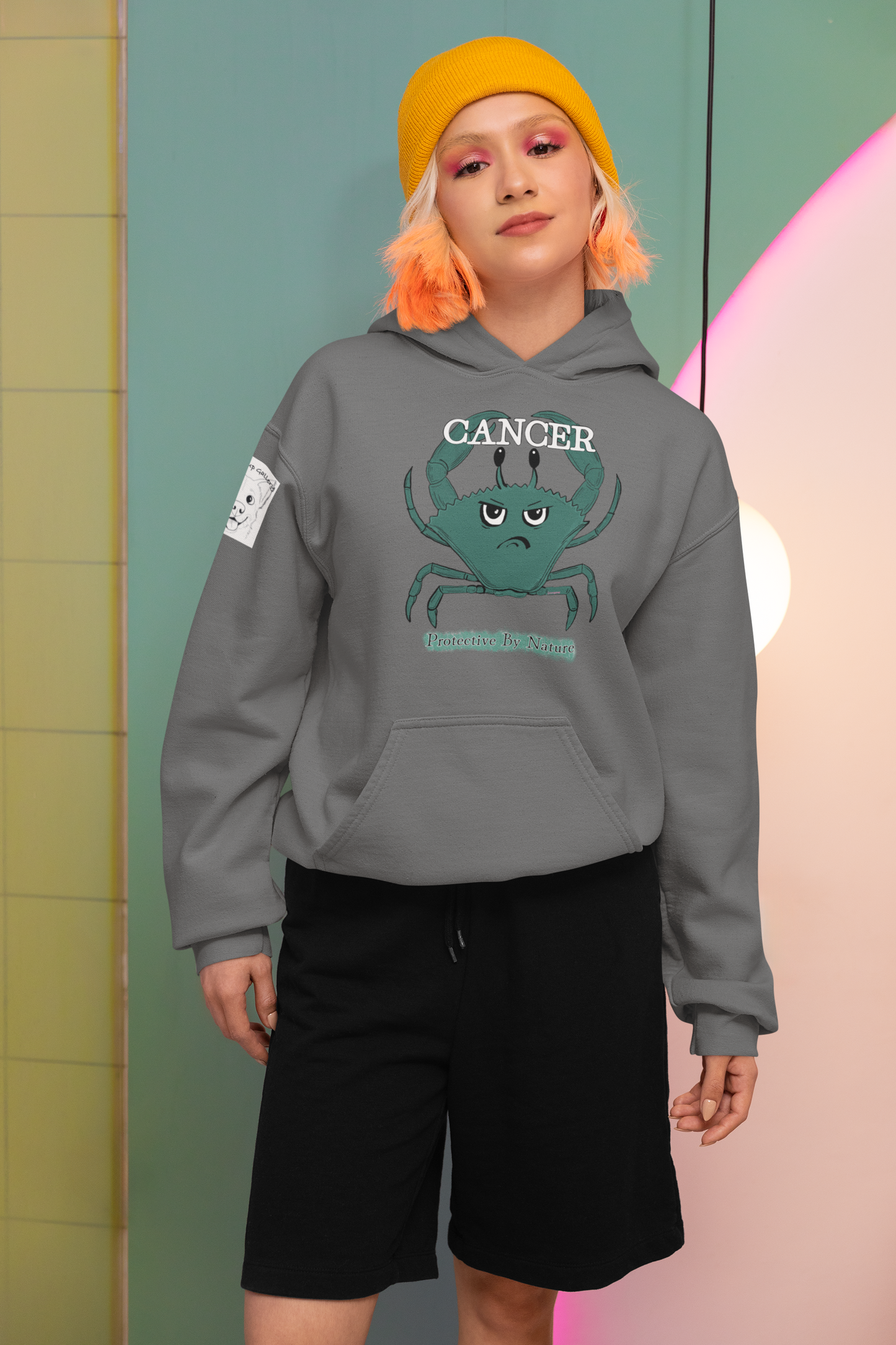 Woman wearing a graphite heather grey Cancer "Protective By Nature" hoodie with black shorts and yellow beanie, posing confidently in a modern colorful room