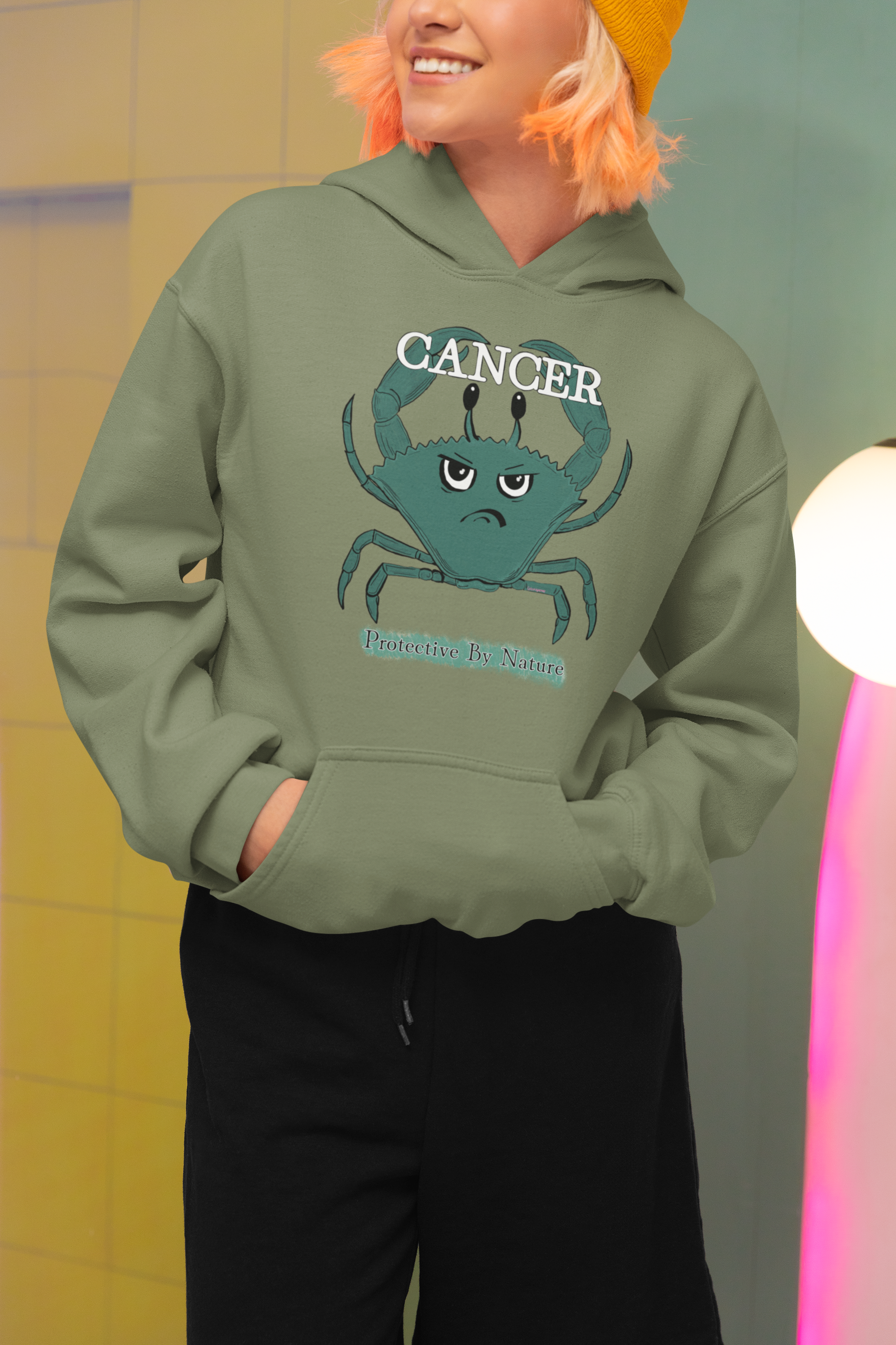 Smiling woman with pink hair wearing an olive military green Cancer "Protective By Nature" hoodie, posing in a vibrant new pop wave inspired setting