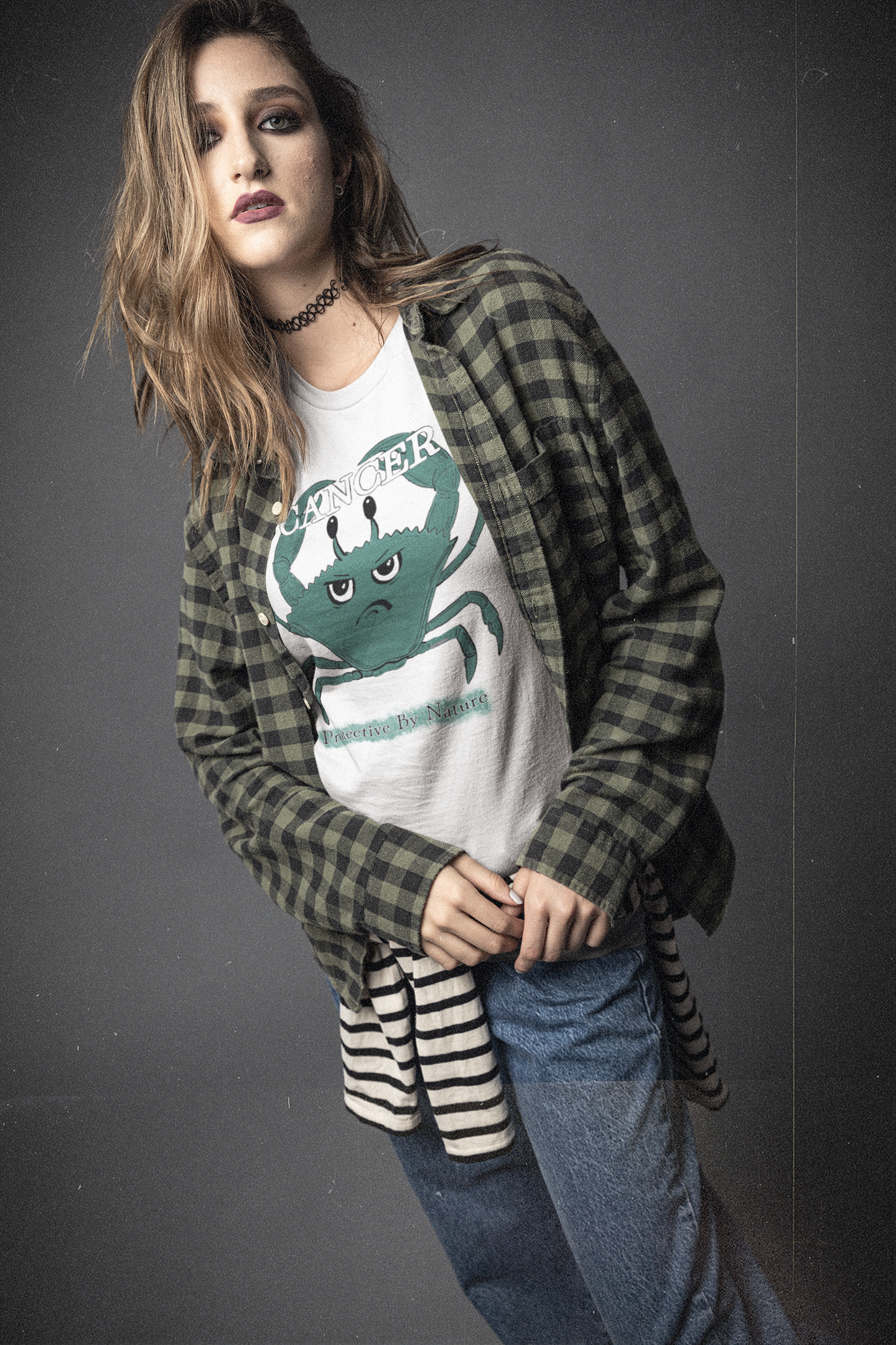 Woman with long blonde hair wearing a white Cancer "Protective By Nature" t-shirt layered under a green and black plaid shirt, standing in a moody studio background