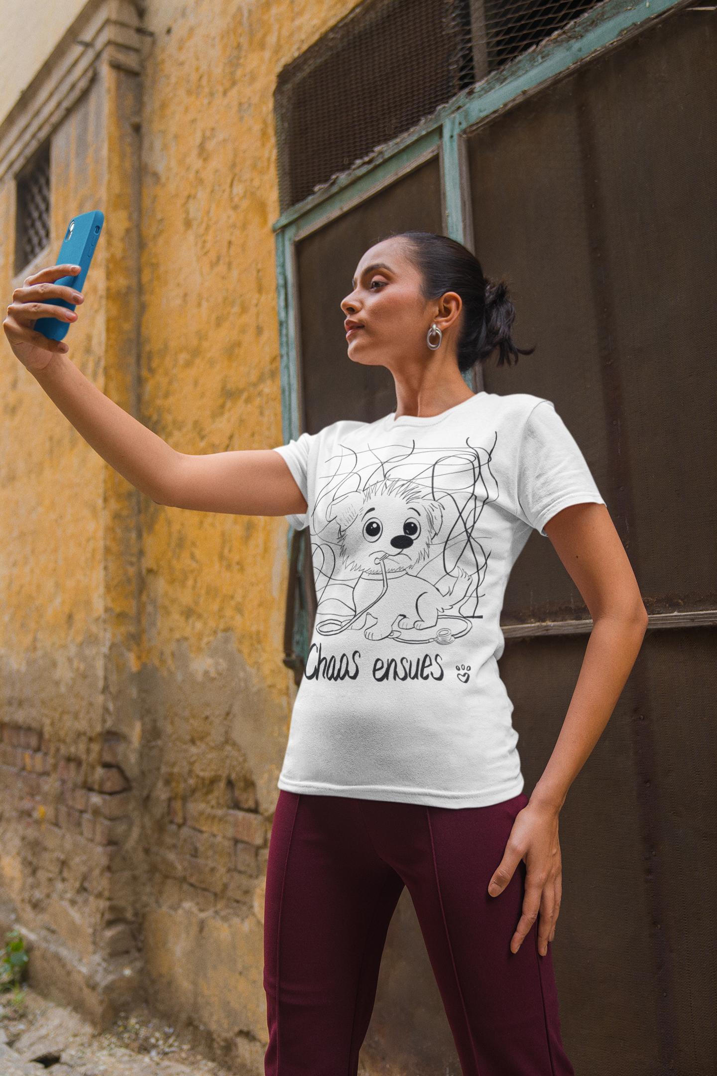 Woman in white Chaos Ensues t-shirt holding up phone for a selfie against old building wall
