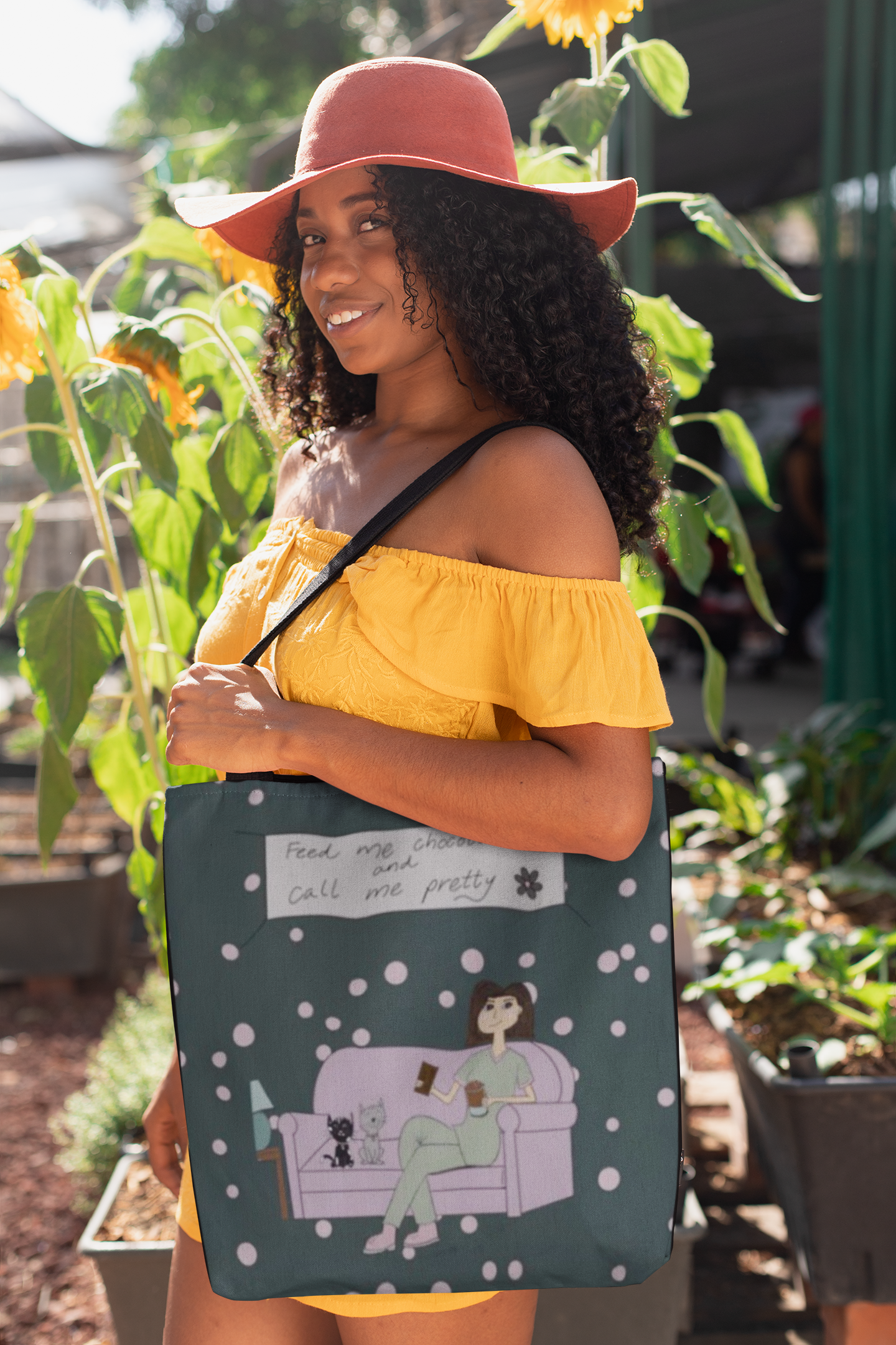 Woman wearing yellow off-shoulder top and sun hat holding a dark green Feed Me Chocolate and Call Me Pretty tote bag outdoors