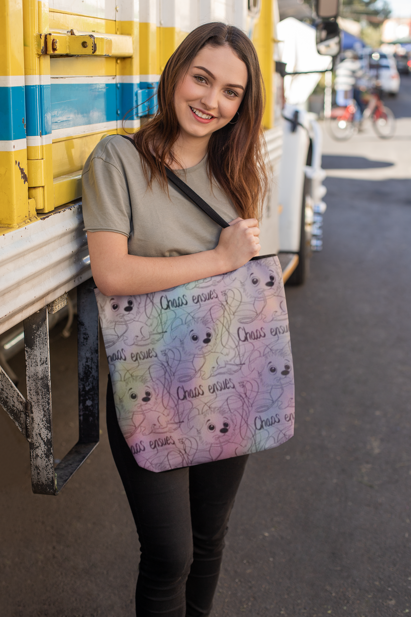 Smiling woman with long brown hair holding a rainbow Chaos Ensues all-over print tote bag in front of a yellow and blue bus
