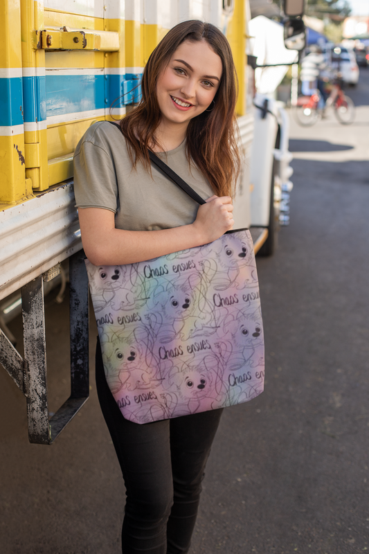 Smiling woman with long brown hair holding a rainbow Chaos Ensues all-over print tote bag in front of a yellow and blue bus