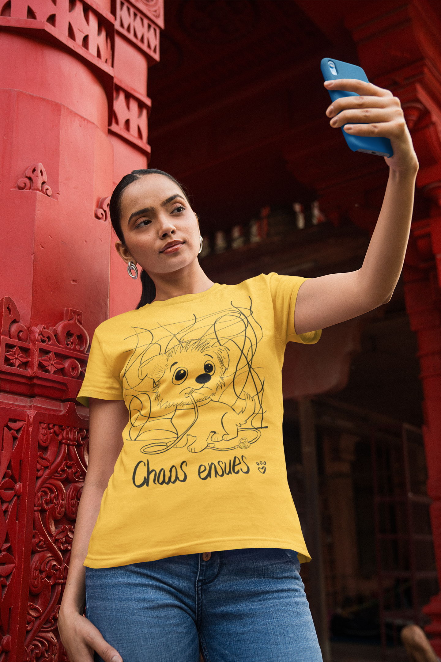 Woman in yellow Chaos Ensues t-shirt taking a selfie in front of ornate red building