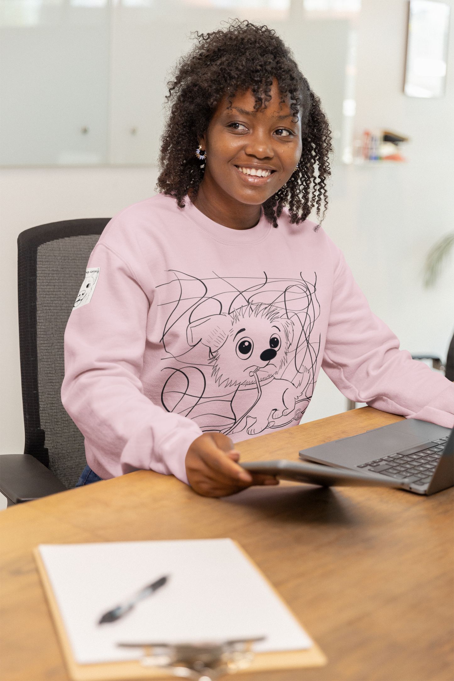 Smiling woman with curly hair wearing a pink Chaos Ensues sweatshirt while working at a desk with laptop