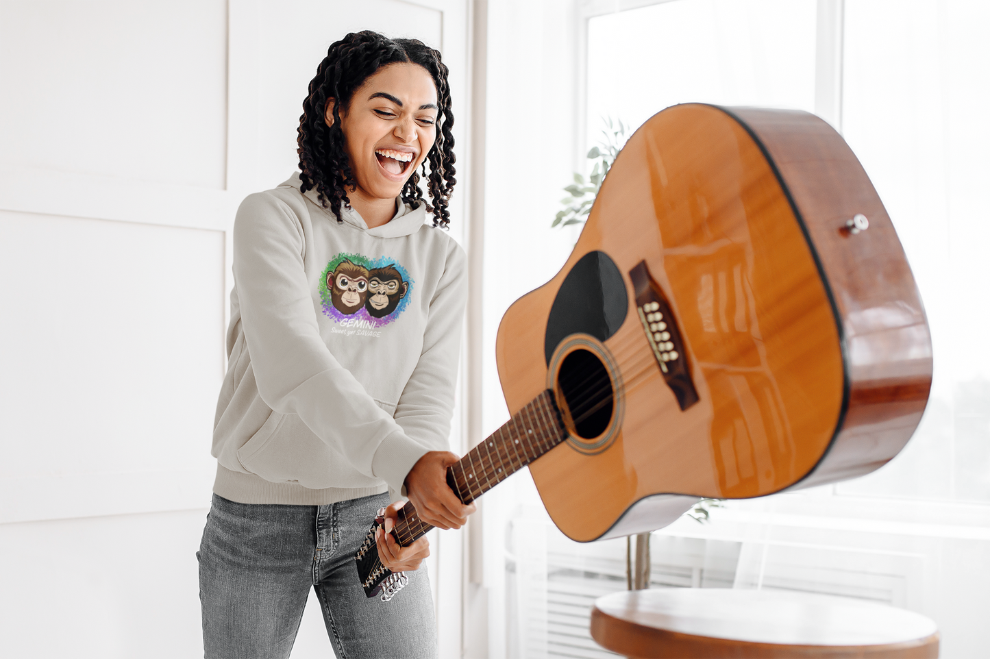Smiling woman with curly hair wearing a carbon grey Gemini "Sweet, yet SAVAGE" hoodie while playfully holding an acoustic guitar in a bright studio