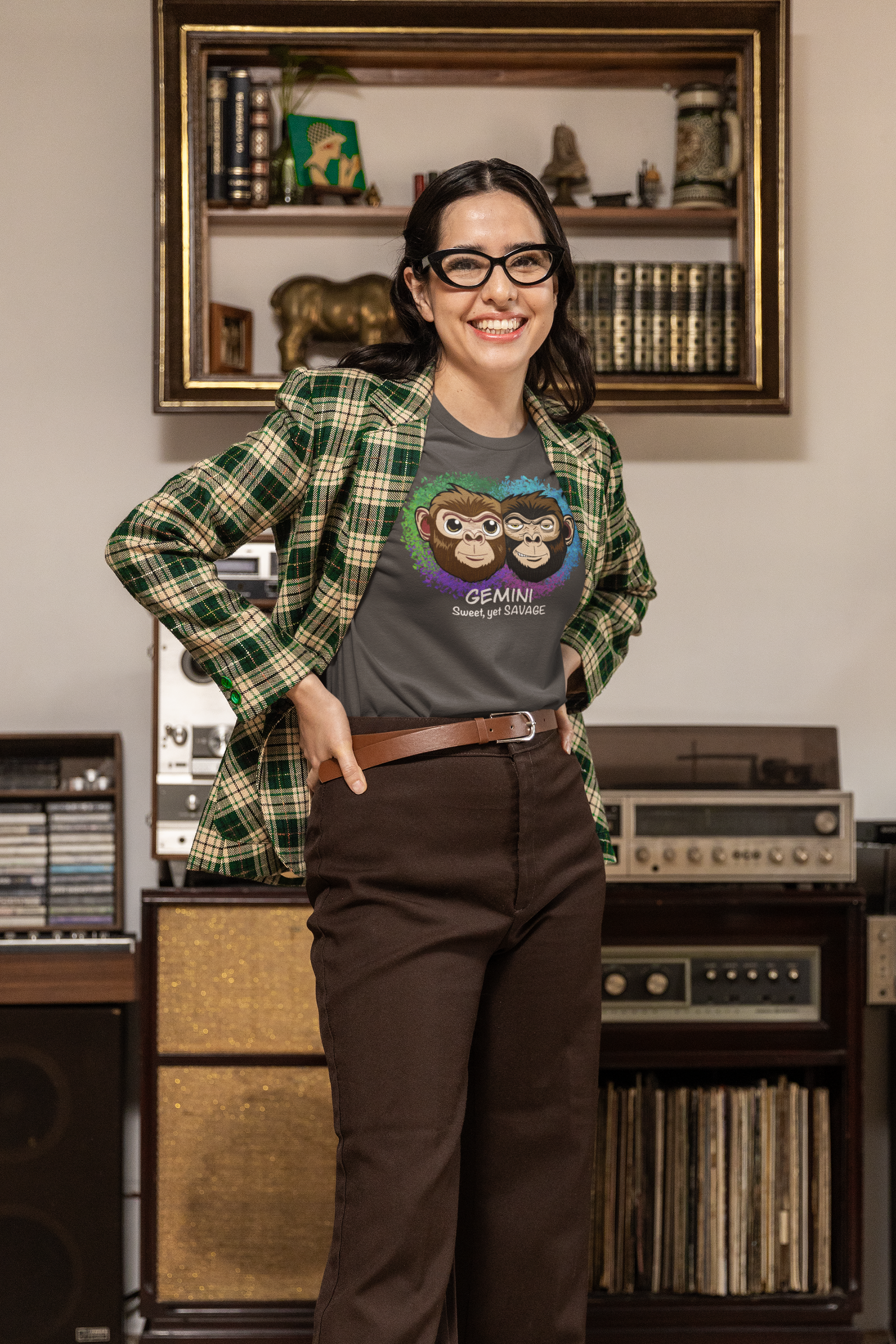Woman with dark hair and glasses wearing a dark heather grey Gemini "Sweet, yet SAVAGE" t-shirt, smiling in a room with vintage decor