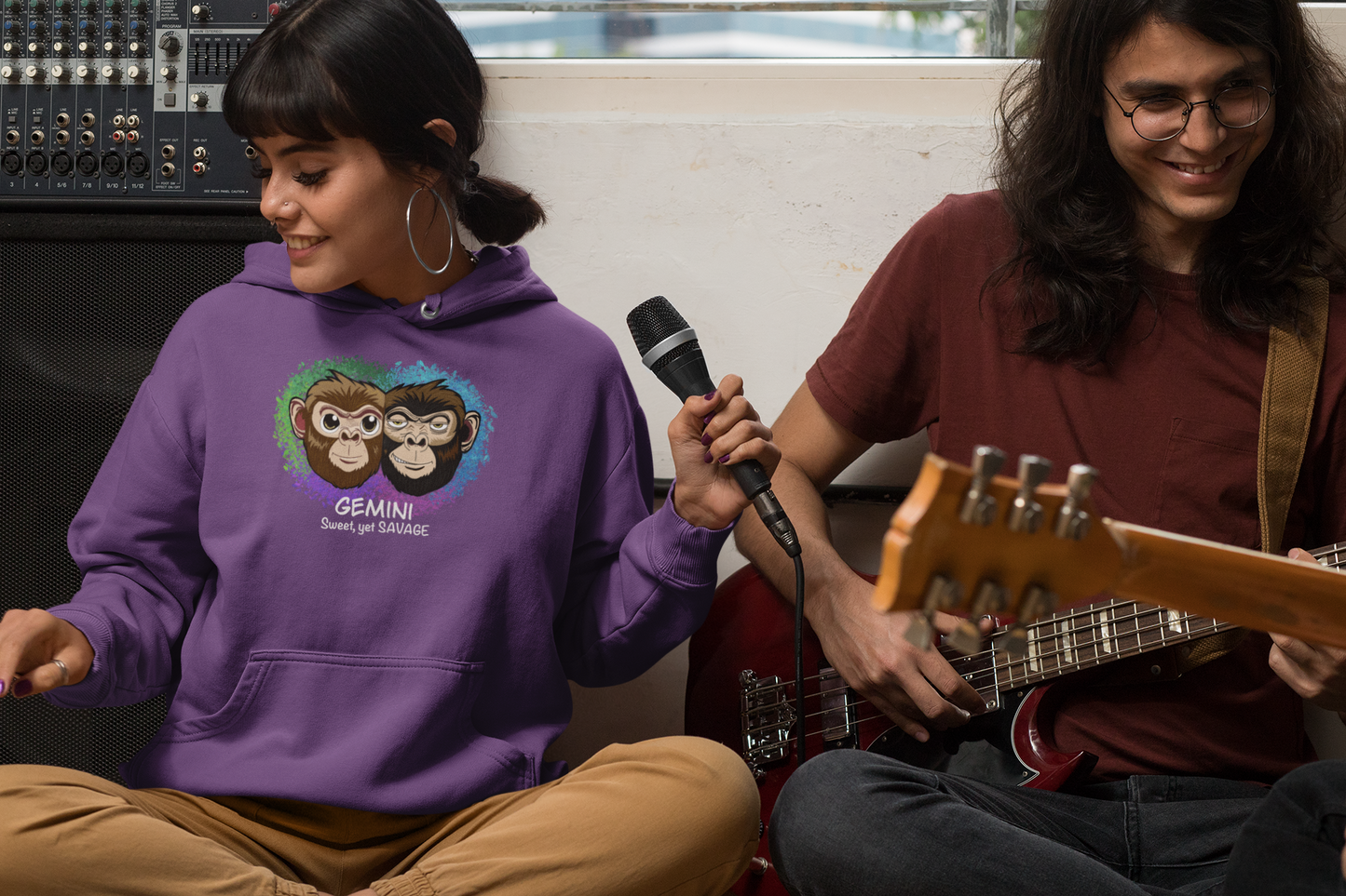 Woman with dark hair and bangs wearing a purple Gemini "Sweet, yet SAVAGE" hoodie holding a microphone next to a man playing guitar