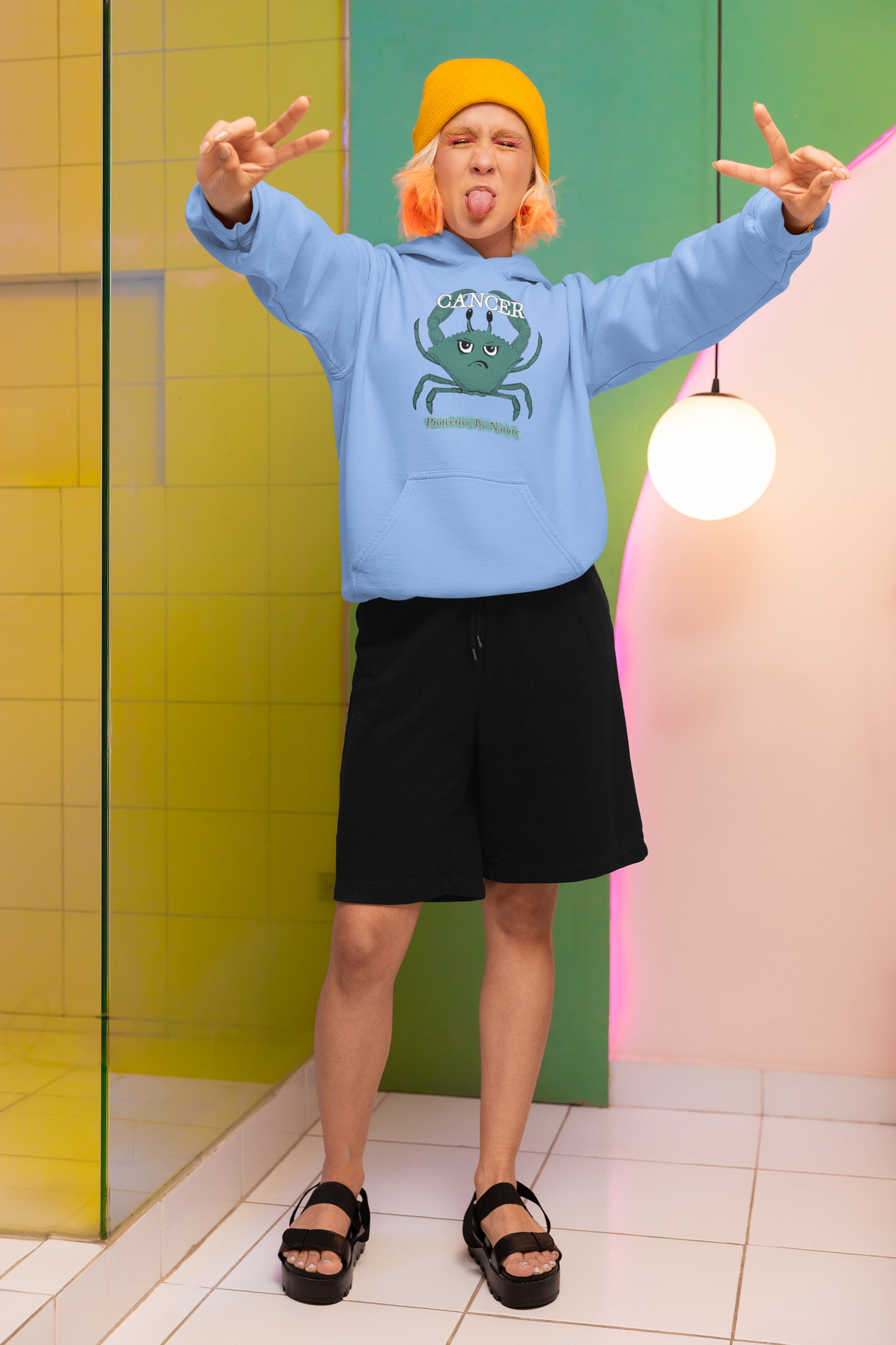 Woman with bright pink hair and yellow beanie wearing a Carolina blue Cancer "Protective By Nature" hoodie, playfully sticking out her tongue in a colorful studio setting