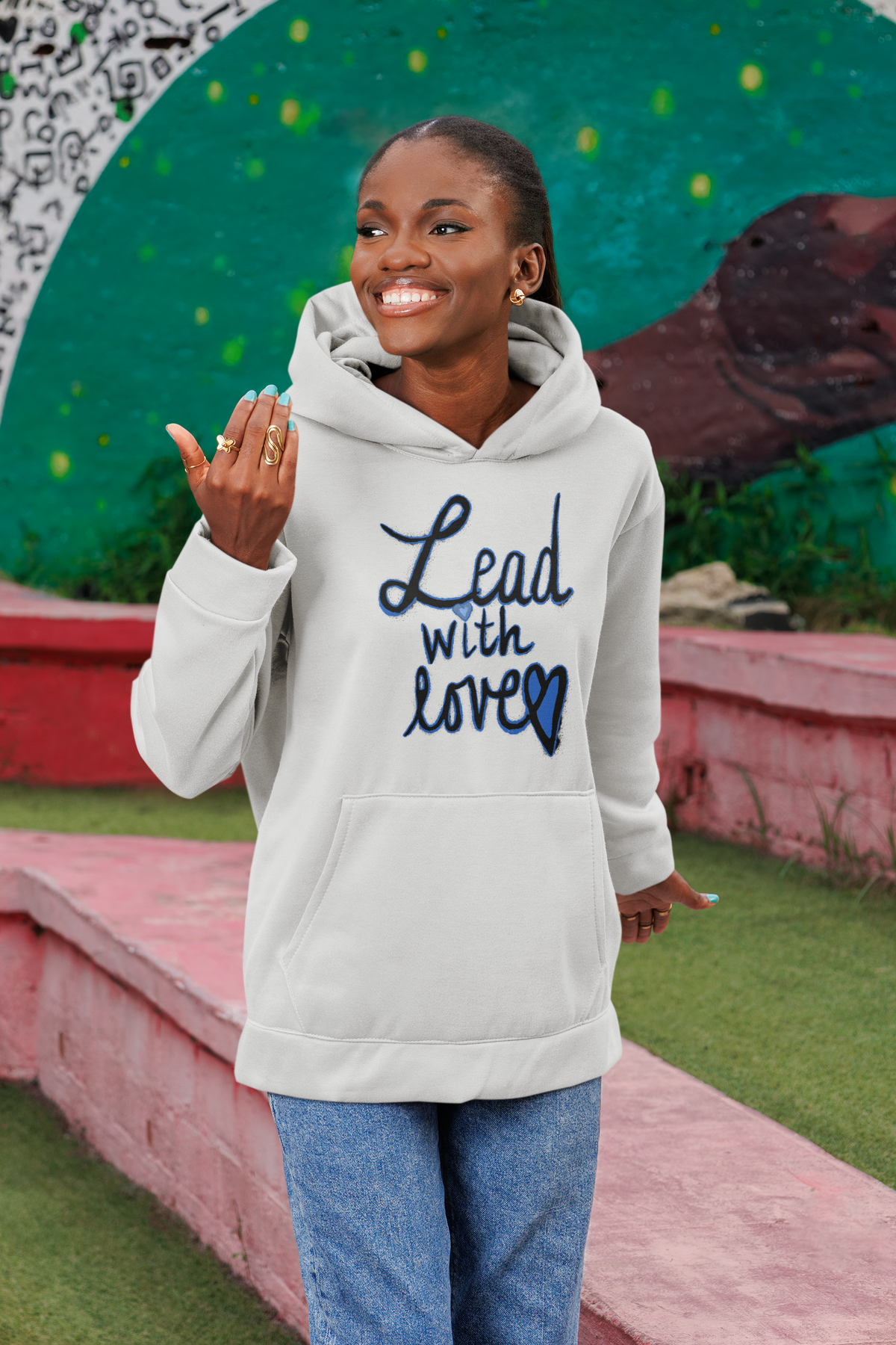 Cheerful woman in an ash grey Lead with love hoodie and jeans gestures while standing in front of a green mural and pink park wall.