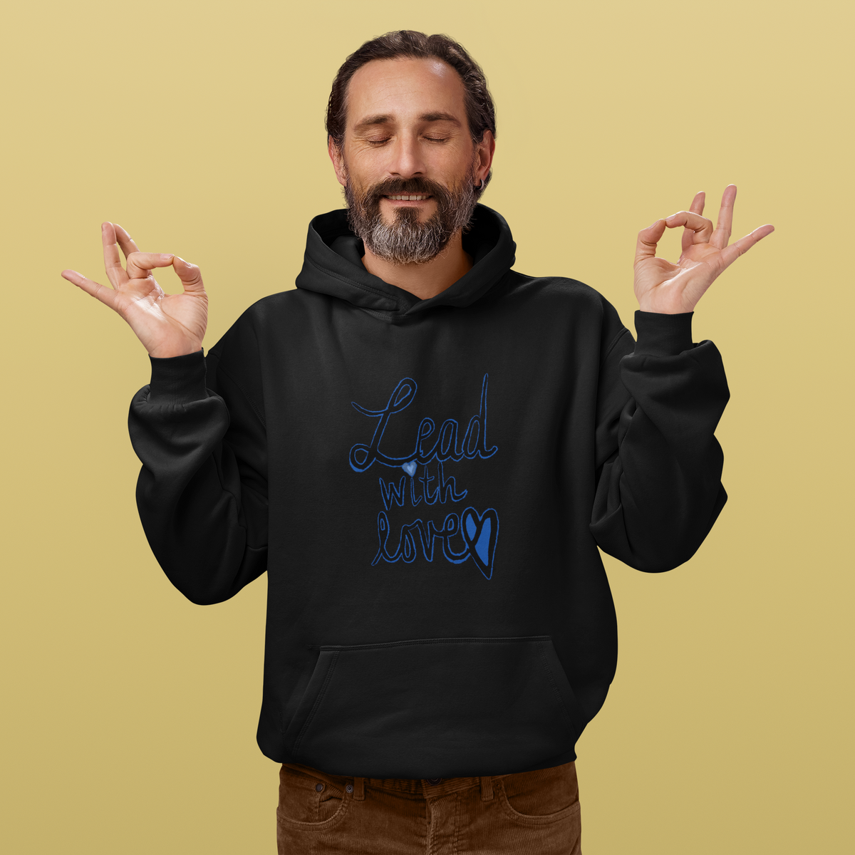 Smiling bearded man with eyes closed holds both hands in a meditation pose, wearing a black Lead with love hoodie and brown cords, on a warm beige background.