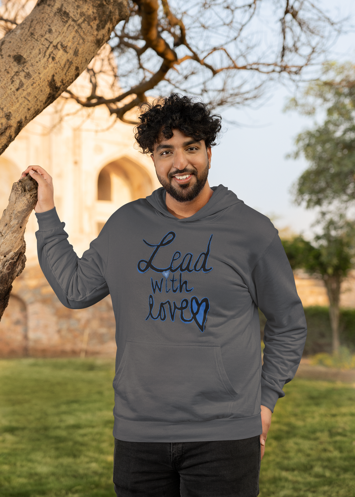 Smiling bearded man in a dark heather Lead with love hoodie rests one hand on a tree trunk, lawn and stone building behind him.