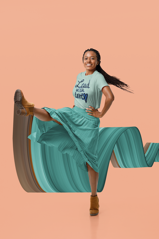 Smiling woman with long braids kicks one leg up in a heather prism dusty blue Lead with love tee, pleated teal skirt, and tan boots.