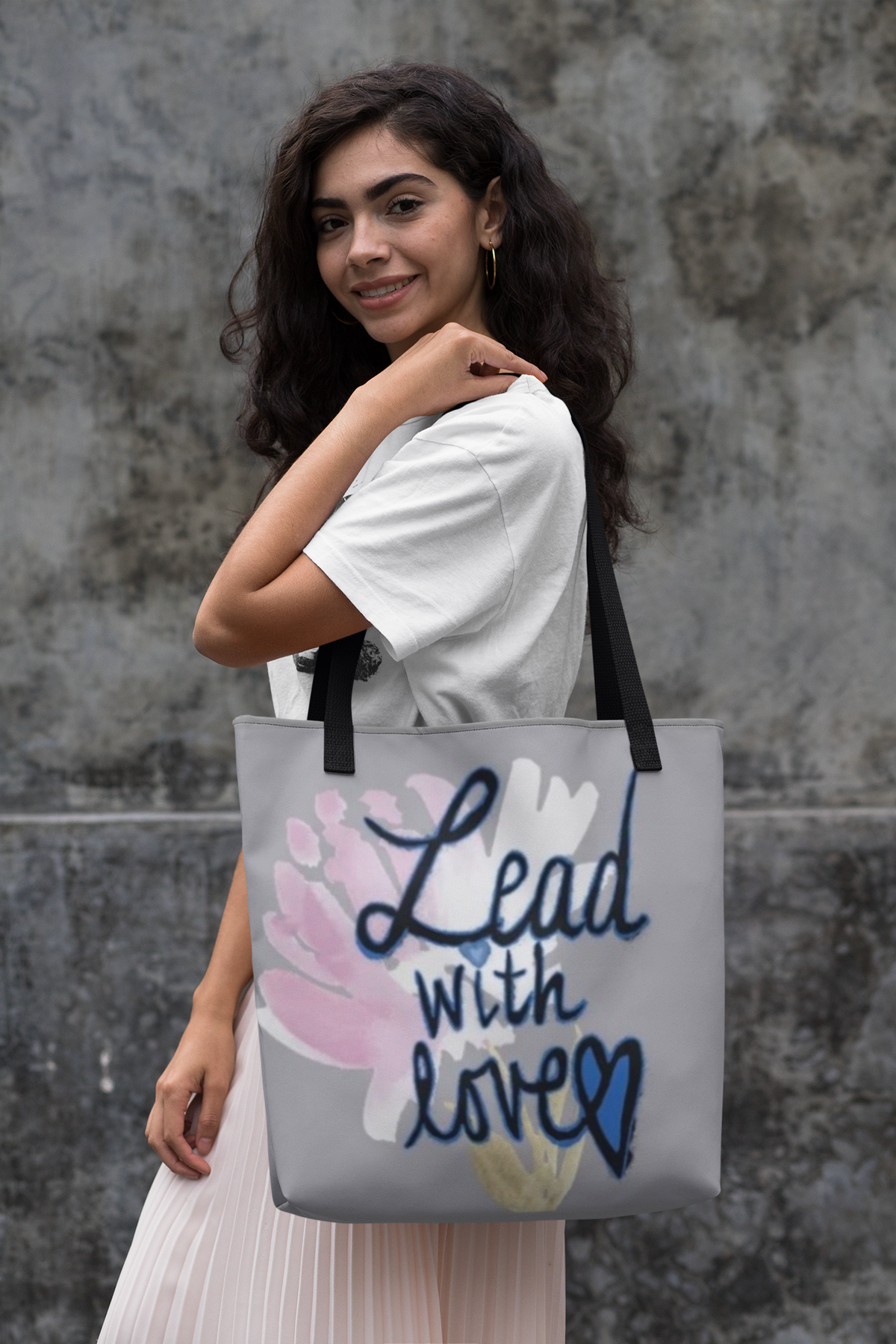 Woman with long dark wavy hair in a white tee and pale pink skirt looks back over her shoulder, grey Lead with Love tote on her arm, against a concrete wall.