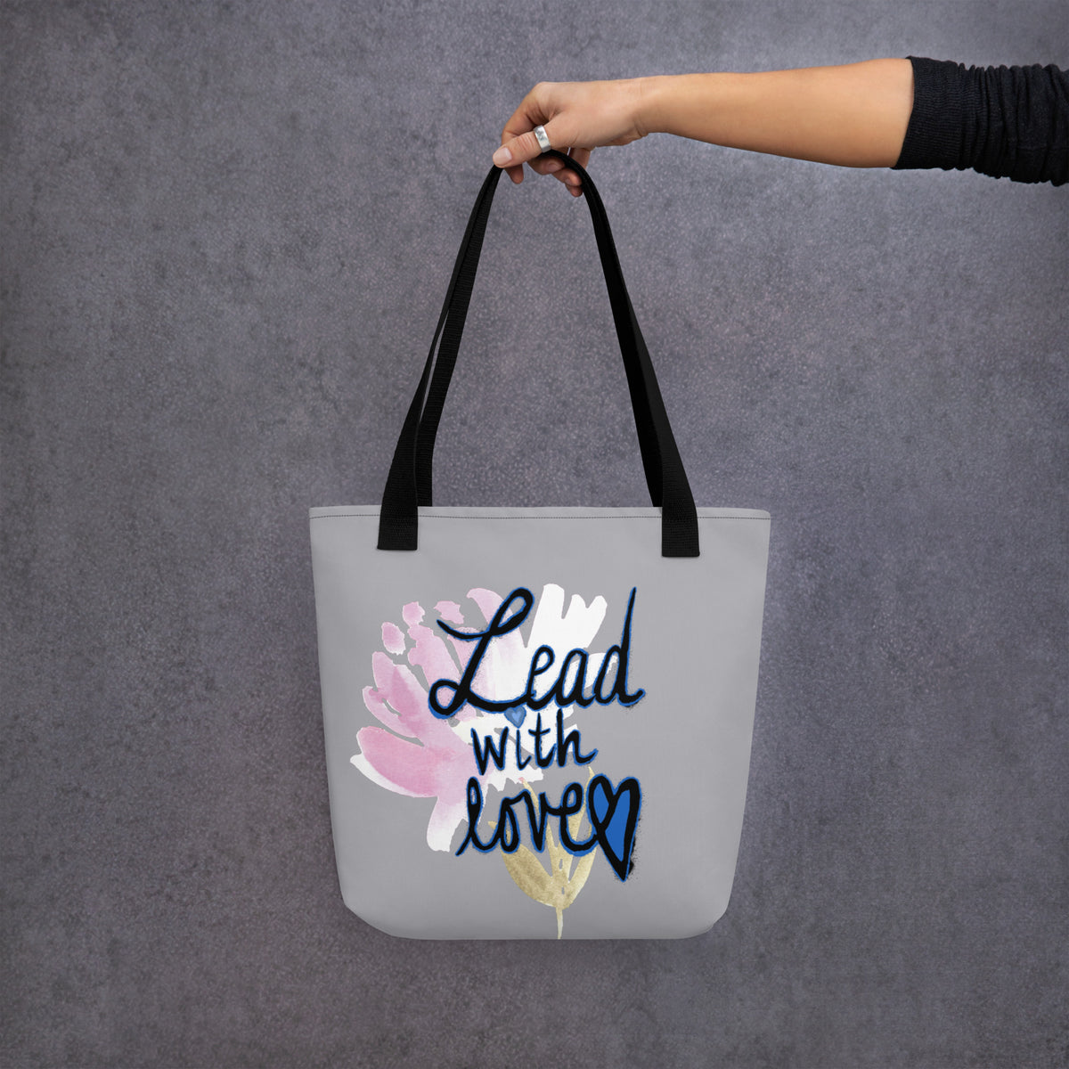 Front view of a grey tote bag with black straps, pink watercolor flower, and navy brush script that reads Lead with love, heart in the last letter, small gold leaf at the bottom.