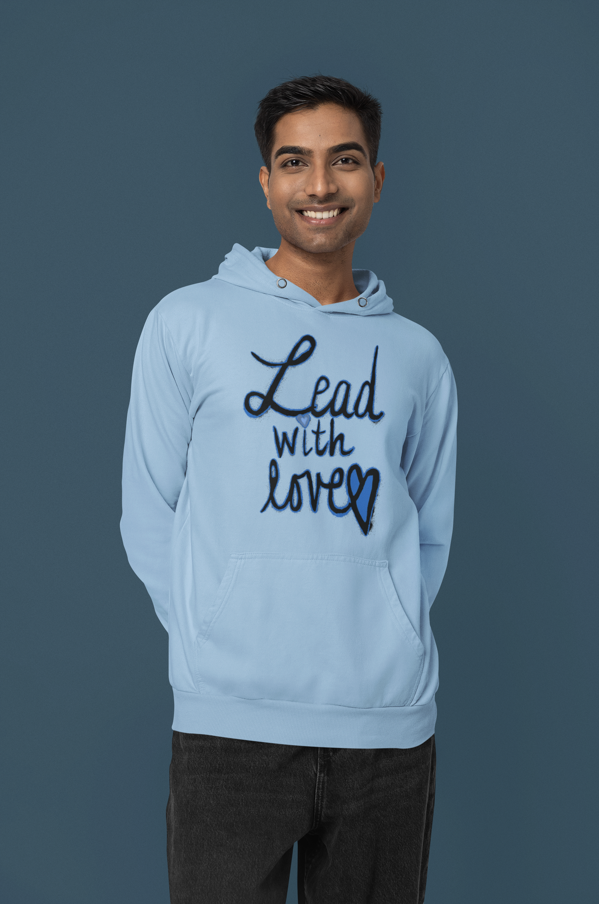 Smiling man in a light blue Lead with love hoodie stands with his hands behind his back against a dark blue studio backdrop.