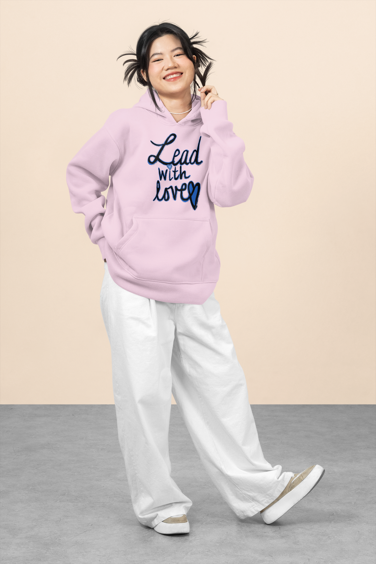 Smiling young woman in a light pink Lead with love hoodie and white wide-leg pants crosses one foot, one hand in the pocket, on a pale studio set.
