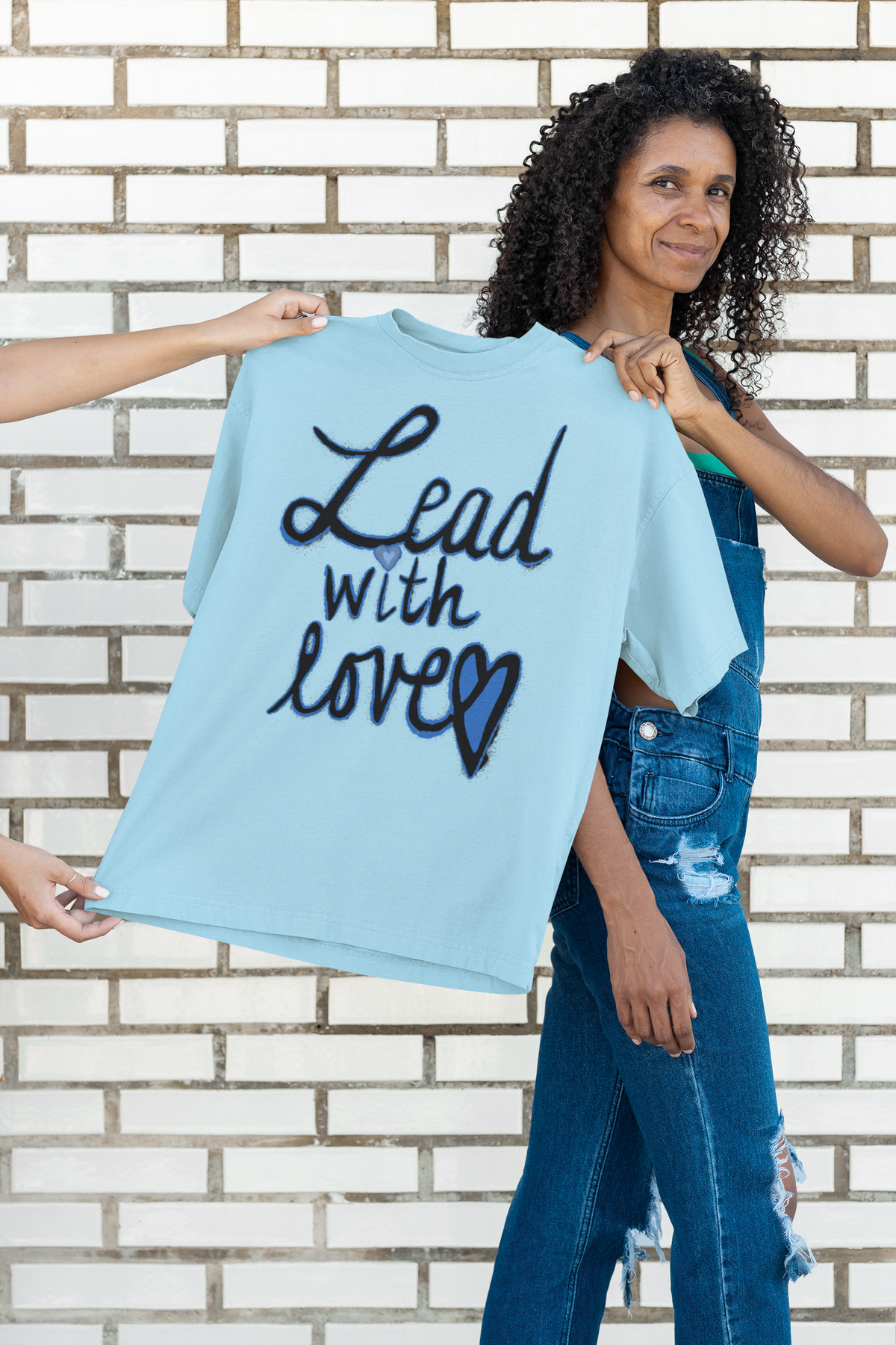 Smiling woman with curly hair in denim overalls holds up an ocean blue tee printed with Lead with love, against a white brick wall.