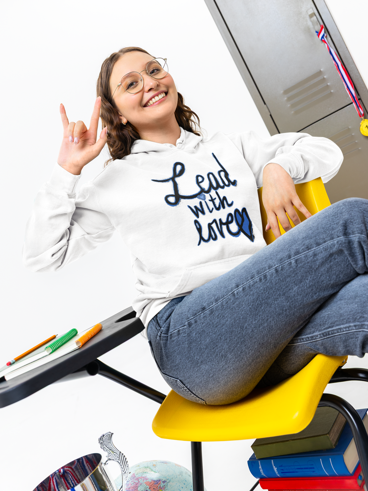 Smiling student with glasses in a white Lead with love hoodie sits on a yellow classroom chair, one hand raised, locker and books behind her.