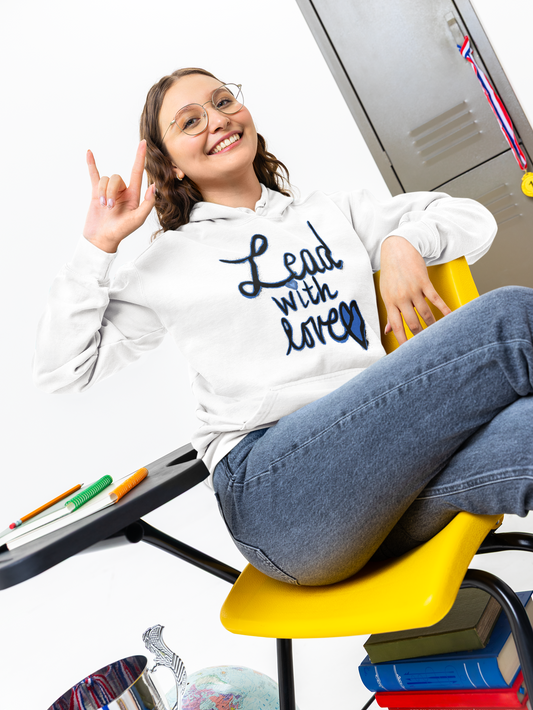 Smiling student with glasses in a white Lead with love hoodie sits on a yellow classroom chair, one hand raised, locker and books behind her.